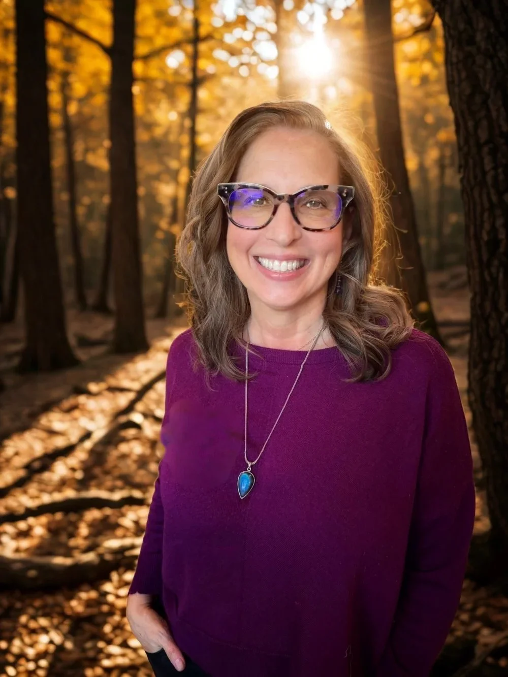 Caryl Rappaport is smiling, wearing glasses and a purple sweater, standing outdoors in a wooded area during sunset.