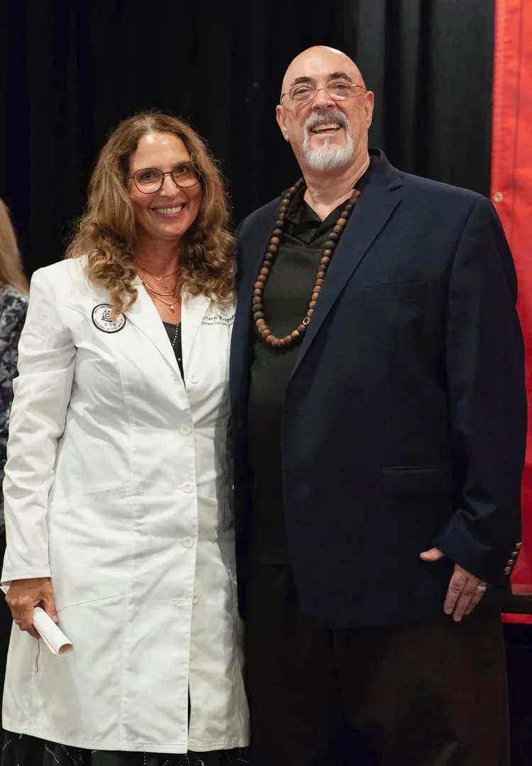 Caryl Rappaport is wearing a white coat and standing with Dr. Jerry Alan Johnson at her graduation ceremony for her Doctorate in Chinese Energetic Medicine.