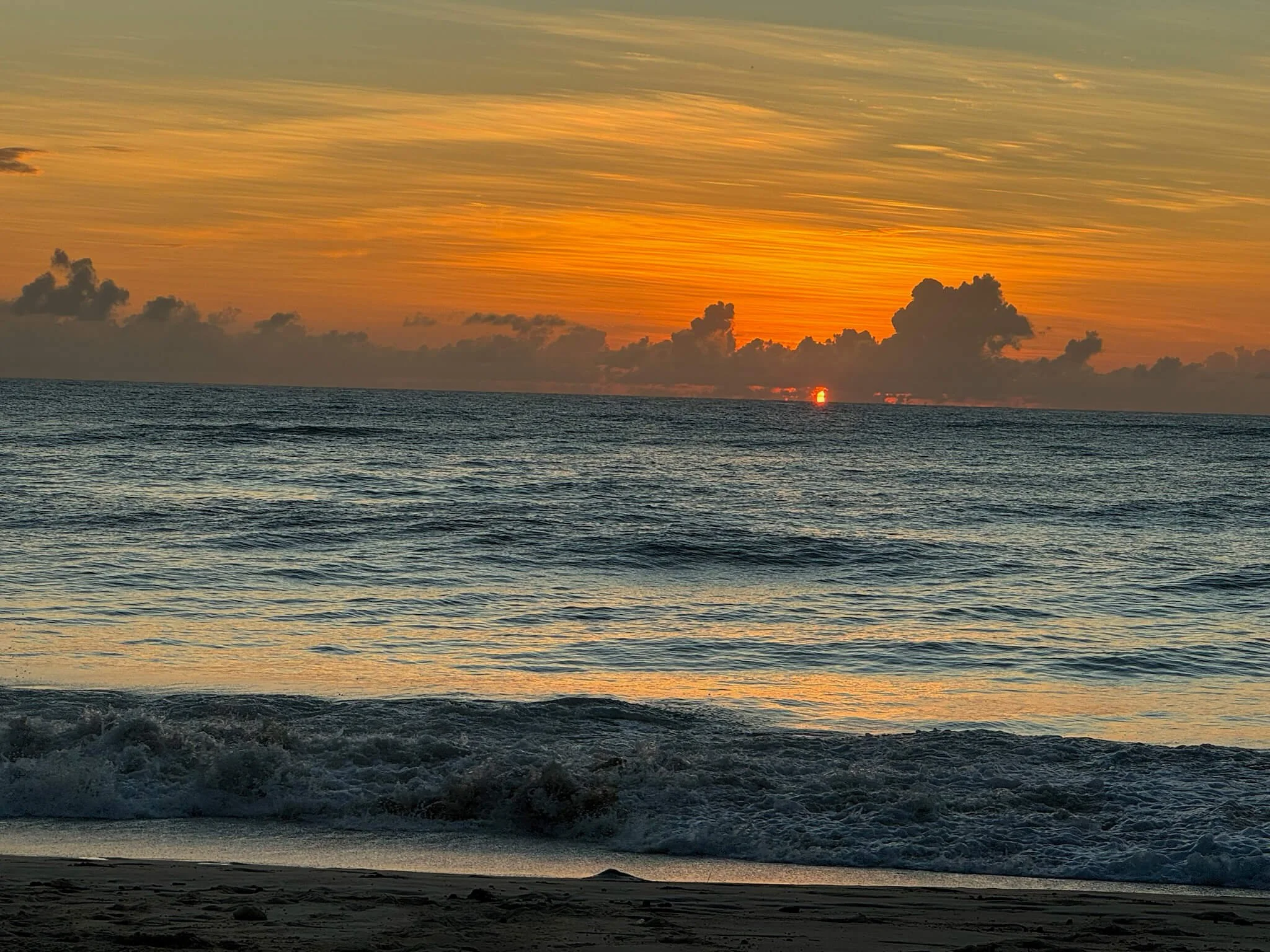 A sunset over the ocean with orange, yellow, and purple hues in the sky, dark clouds near the horizon, and gentle waves in the foreground.