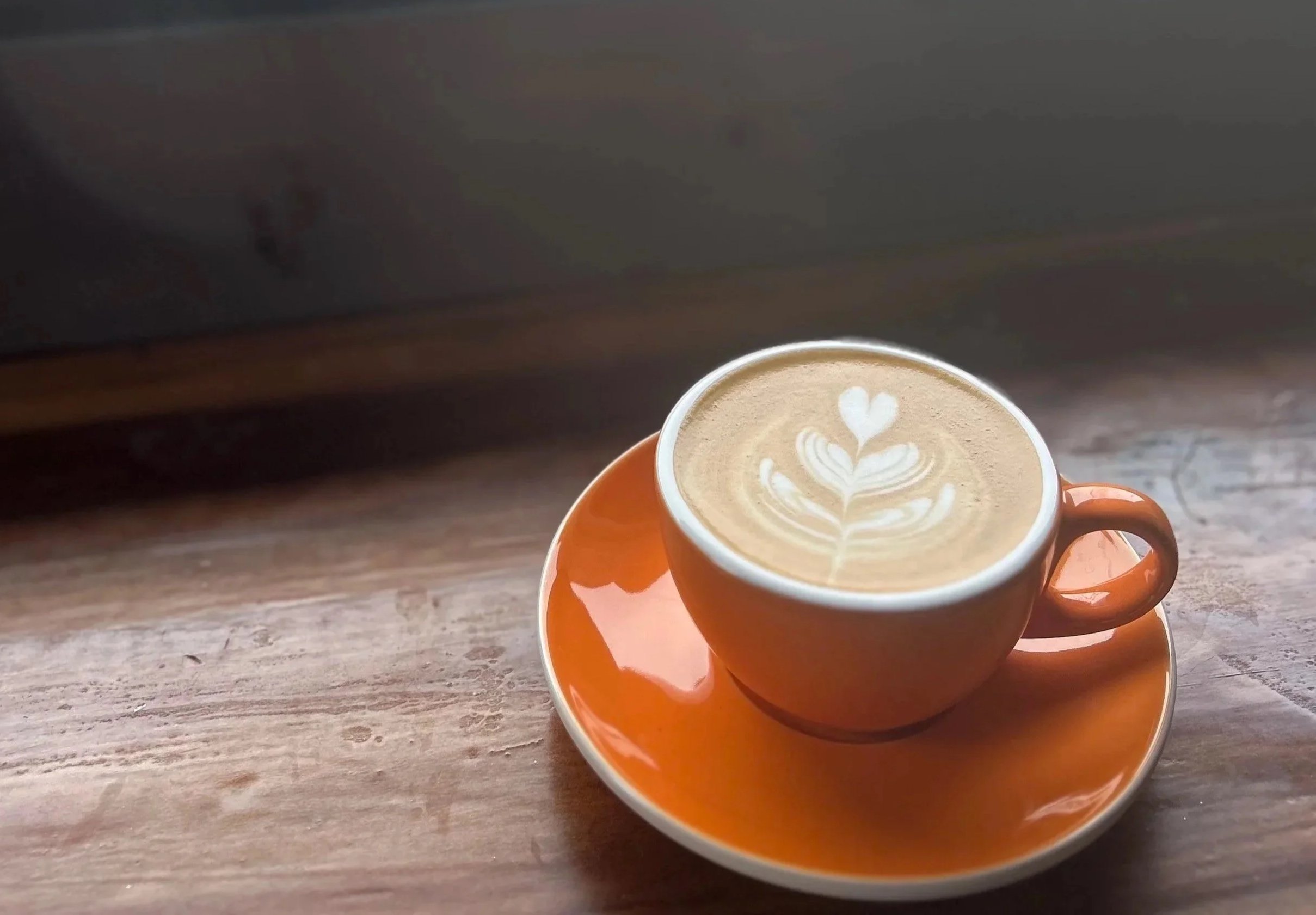 A cup of coffee with latte art on top, placed on an orange saucer on a wooden table.