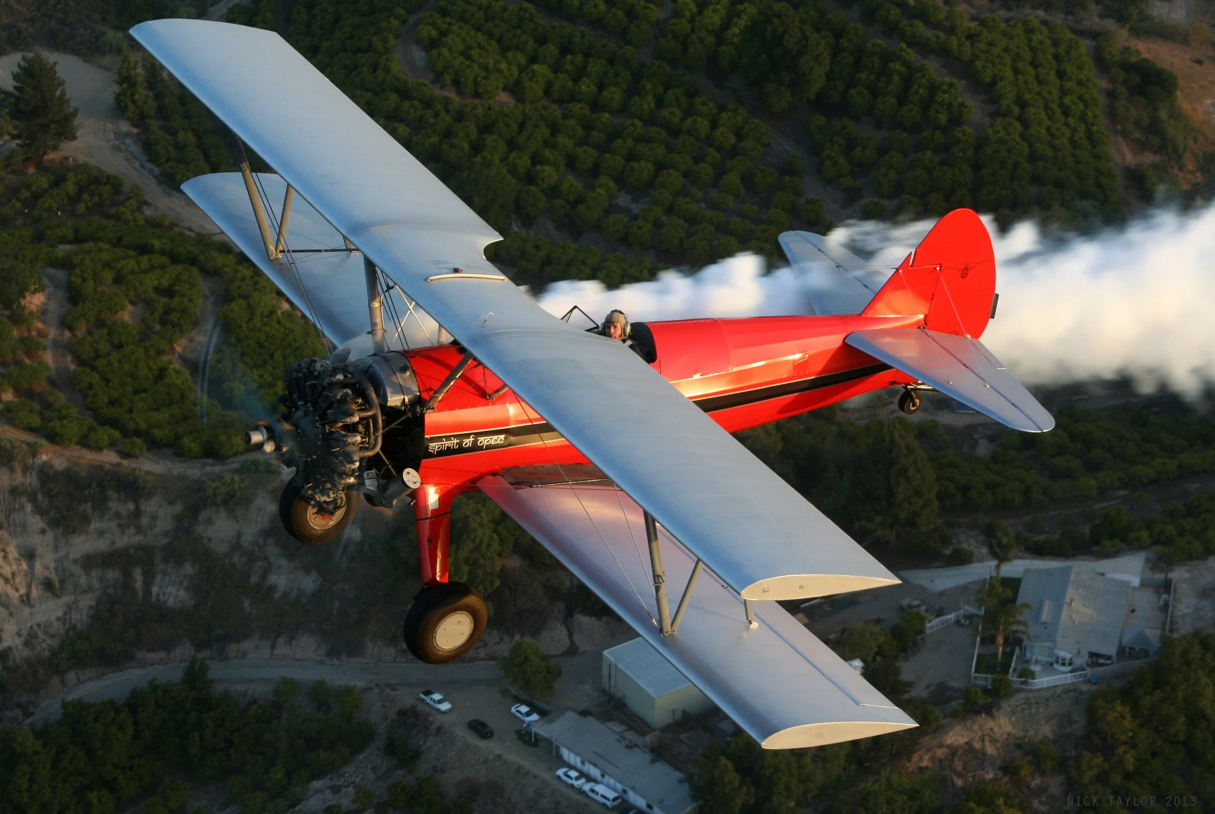 A red vintage biplane flying over a landscape of forests, hills, and buildings with a trail of white smoke behind it.