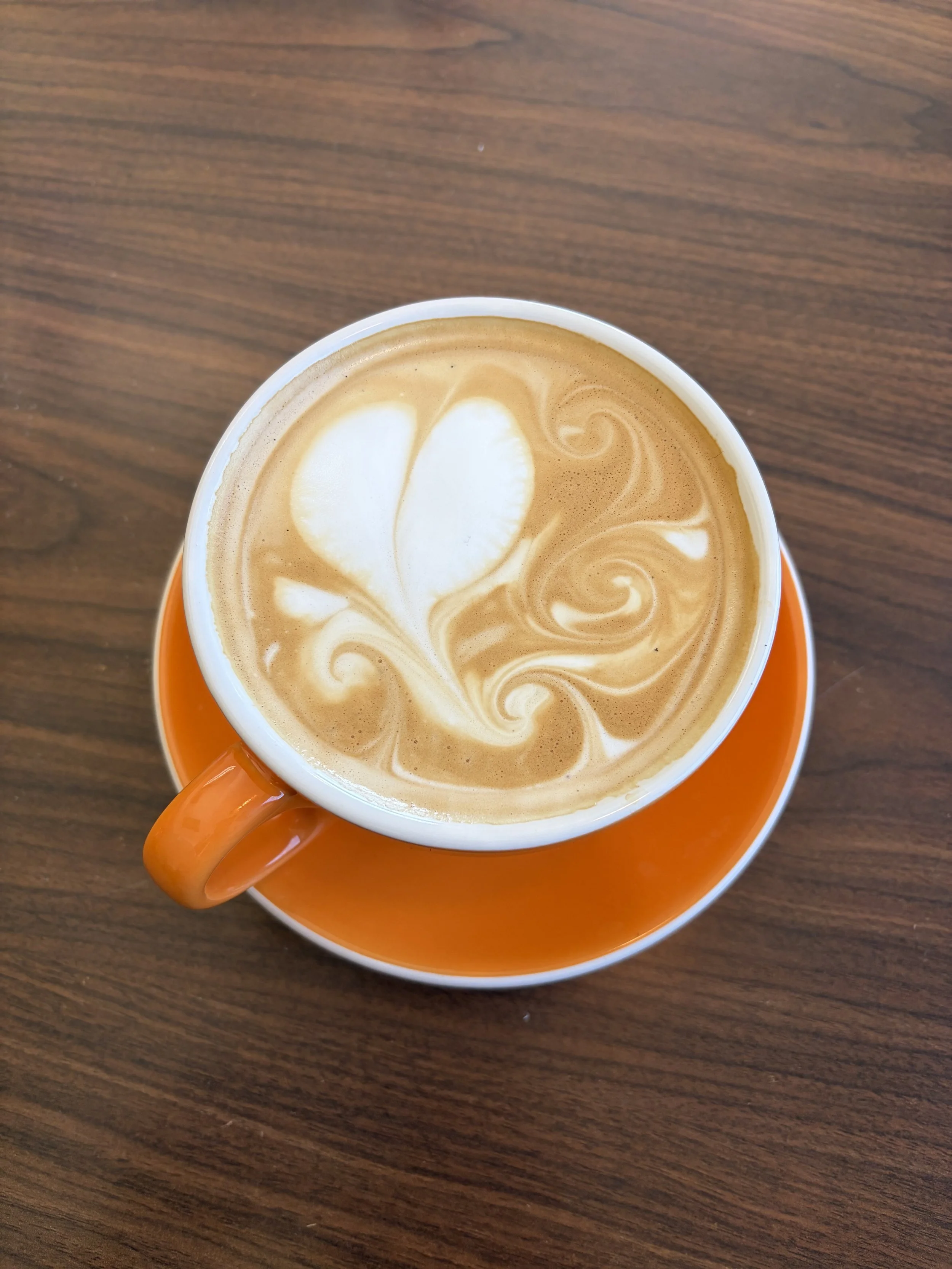 A cup of coffee with latte art on top, placed on an orange saucer, on a wooden table.