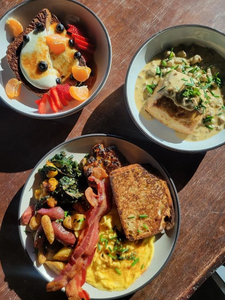 A breakfast spread with three bowls containing different foods: a bowl with French toast topped with fruit, whipped cream, and syrup; a bowl with a piece of fish in a creamy sauce with chopped herbs; and a bowl with scrambled eggs, bacon, sautéed vegetables, and toast.