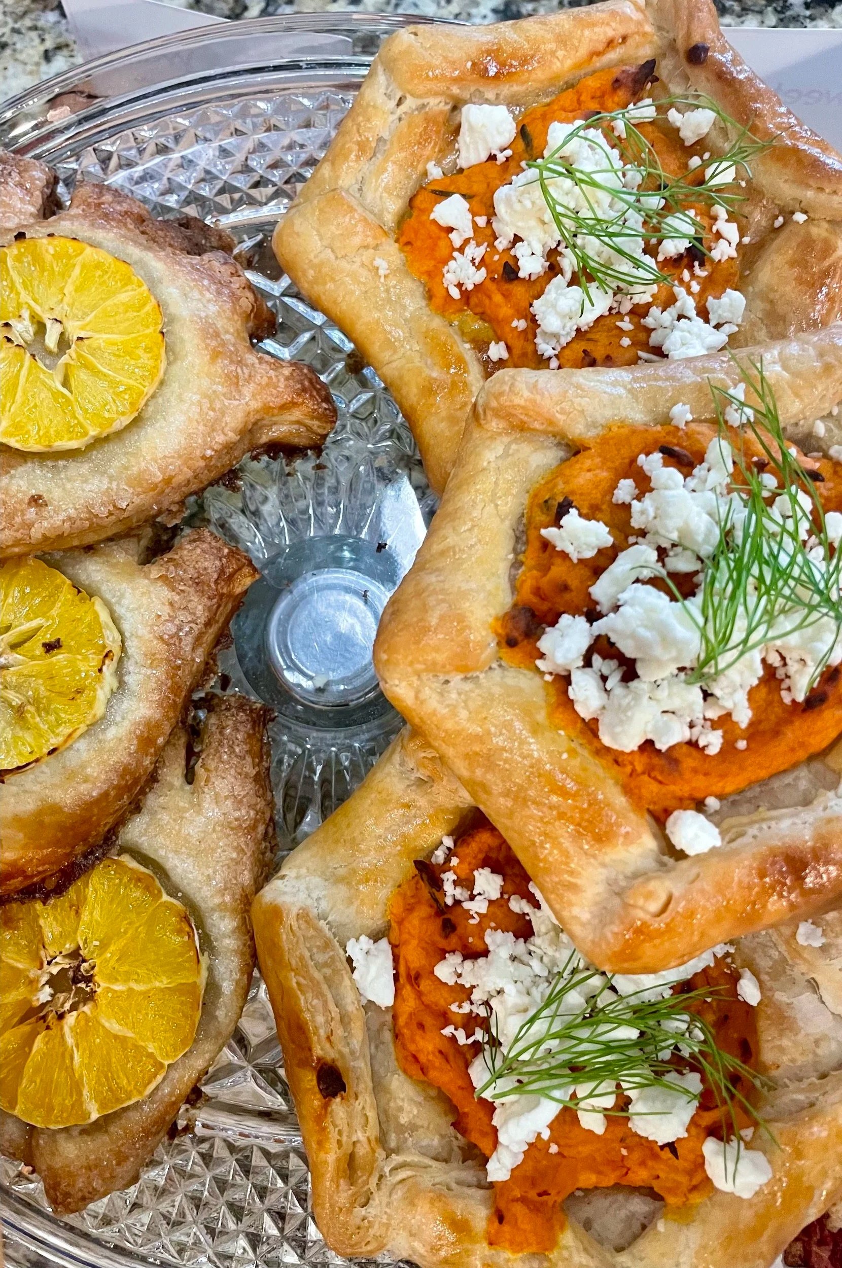 Two pastry squares topped with orange sweet potato mash, crumbled white cheese, and garnished with fresh green herbs, next to two baked slices of bread with lemon slices on top, on a glass serving dish.
