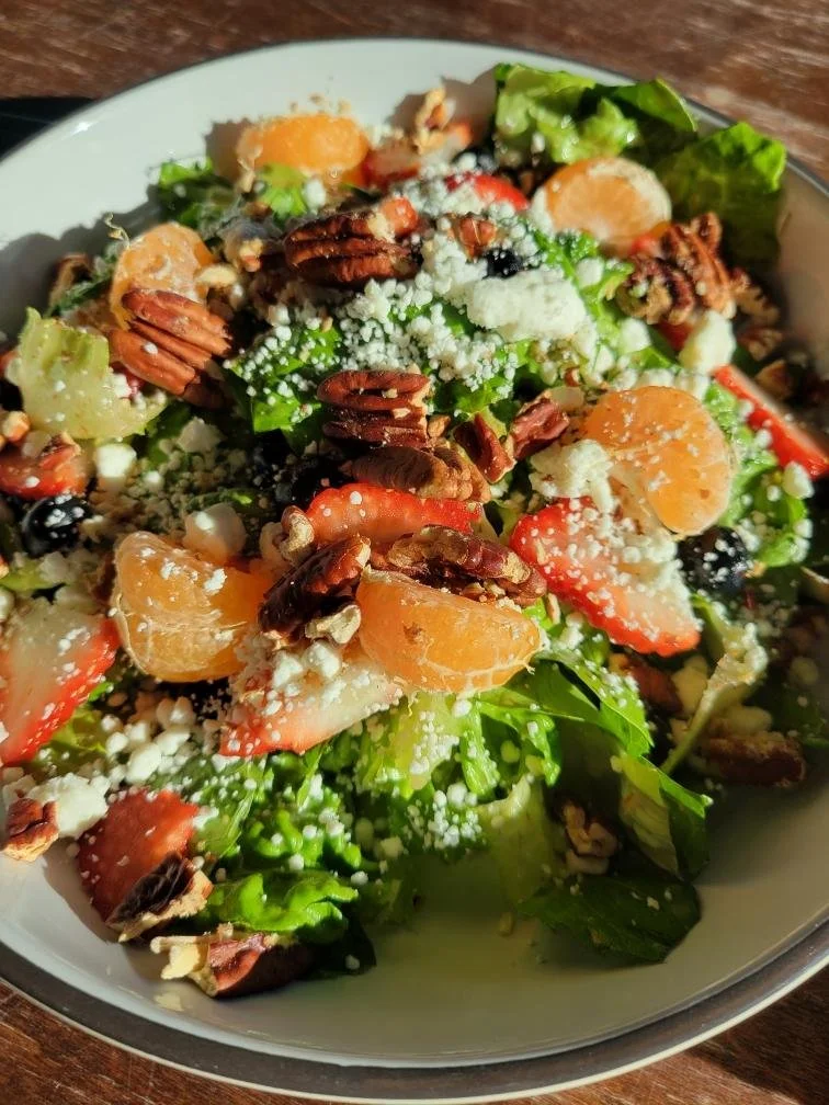 Close-up of a fresh salad with chopped lettuce, strawberries, mandarin orange segments, pecans, crumbled cheese, and black olives in a white bowl on a wooden surface.