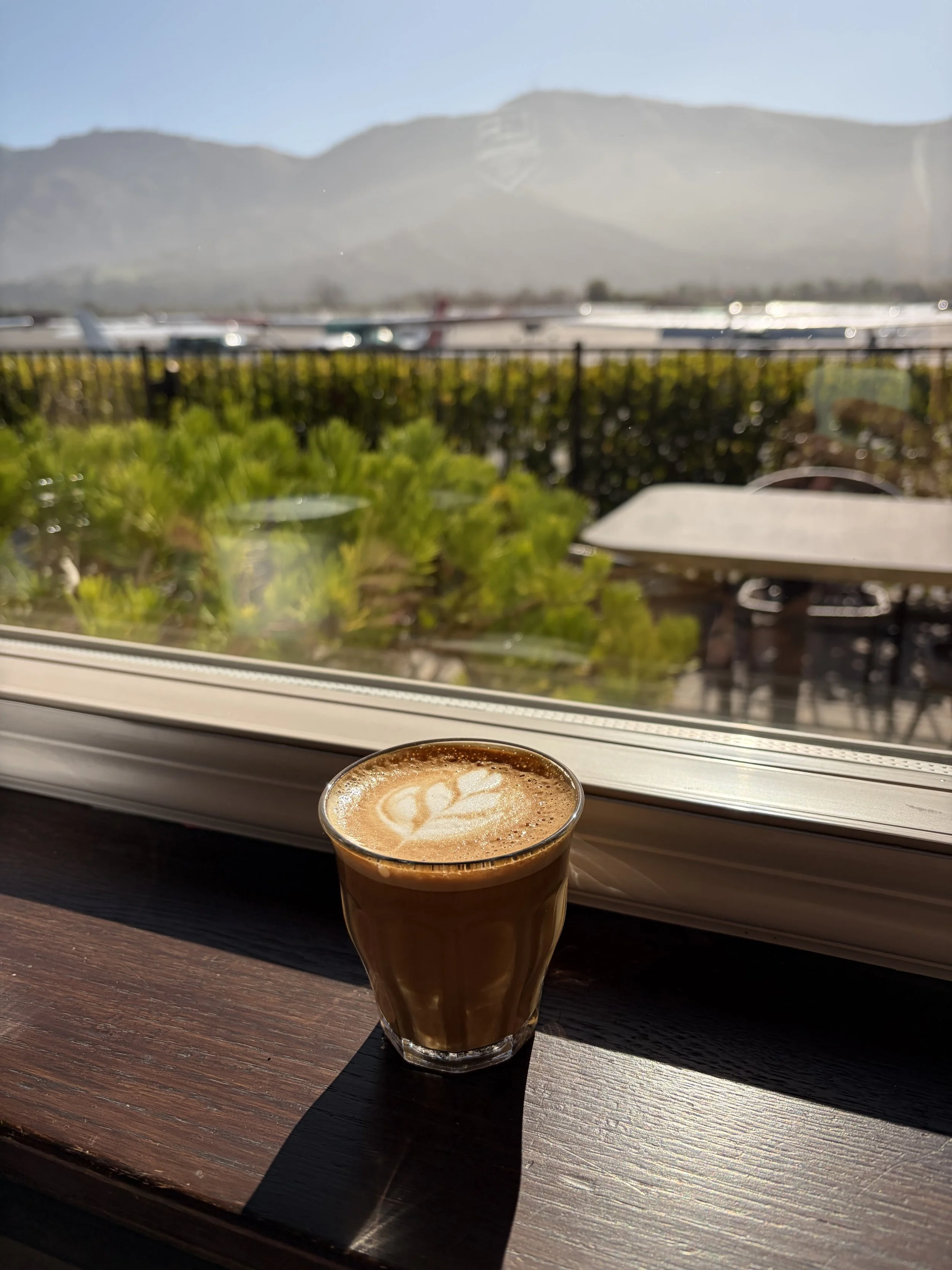 A glass of coffee with latte art on top sits on a wooden windowsill, with a view of the outdoor patio, greenery, and mountains in the distance.