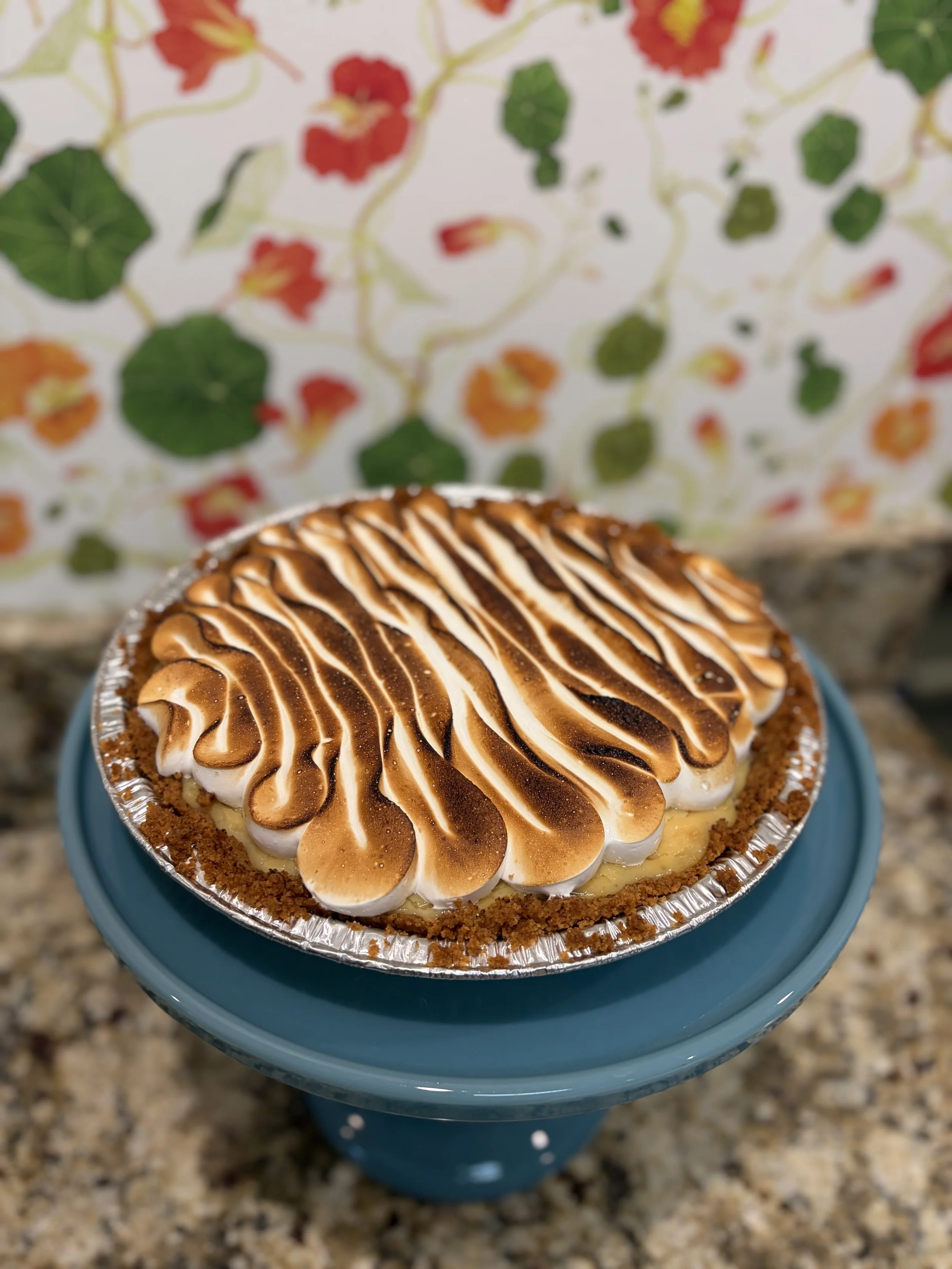 A baked cheesecake topped with toasted marshmallow-like meringue and chocolate sauce, placed on a blue cake stand with a floral wallpaper background.
