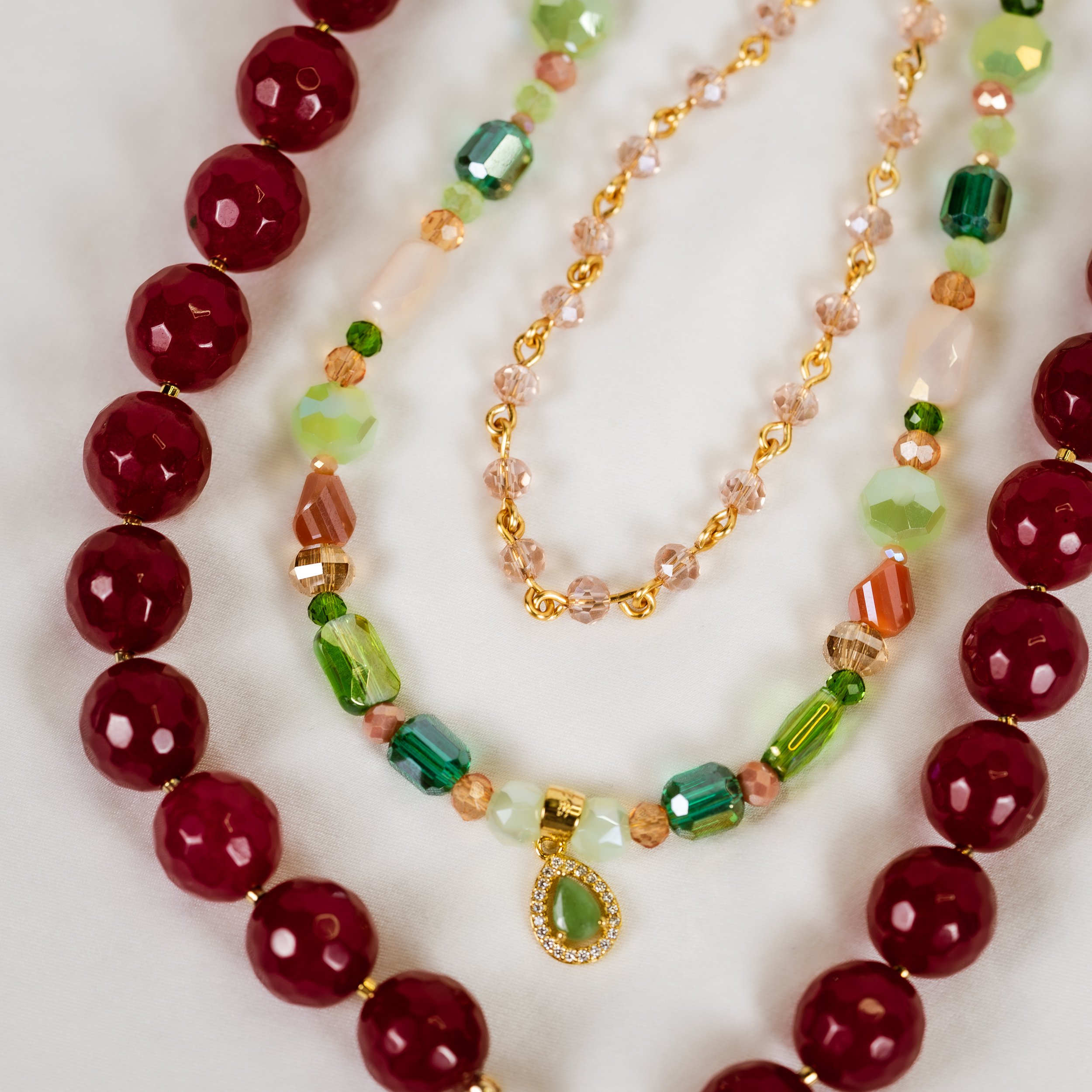 Colorful beaded necklaces on a white surface, including red, green, pink, and orange beads, with one gold chain necklace featuring a teardrop-shaped pendant with green and clear stones.