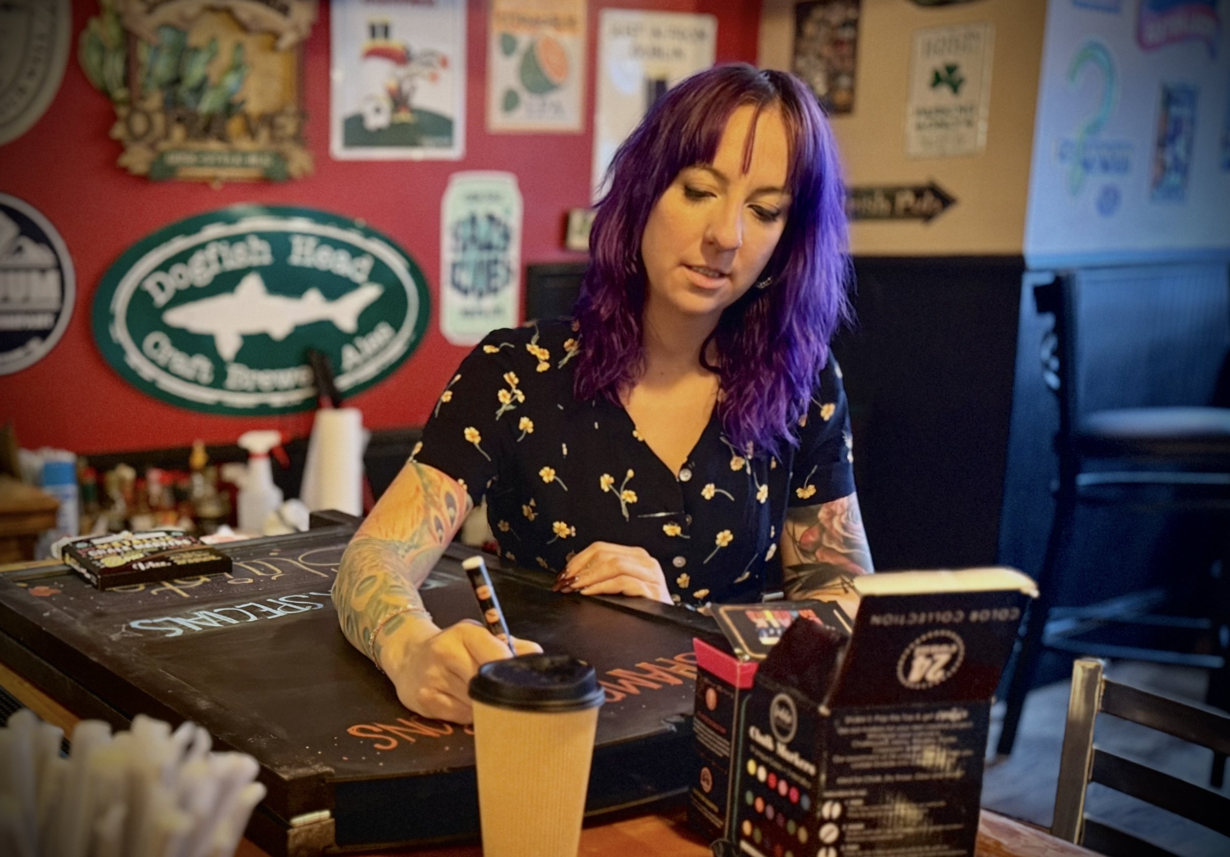 A woman with purple hair and floral tattoos sitting at a bar, writing on a blackboard on the table, with a coffee cup and a menu in front of her, in a colorful, decorated pub or restaurant setting.