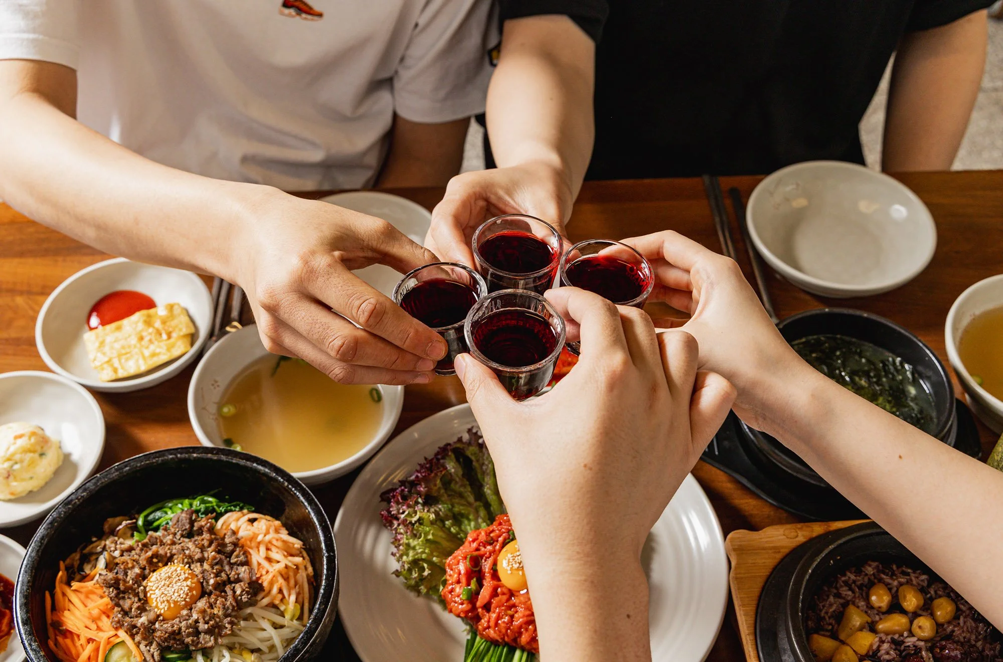People raising glasses of red wine in a toast over a table filled with Korean dishes such as bibimbap, side dishes, and soup.