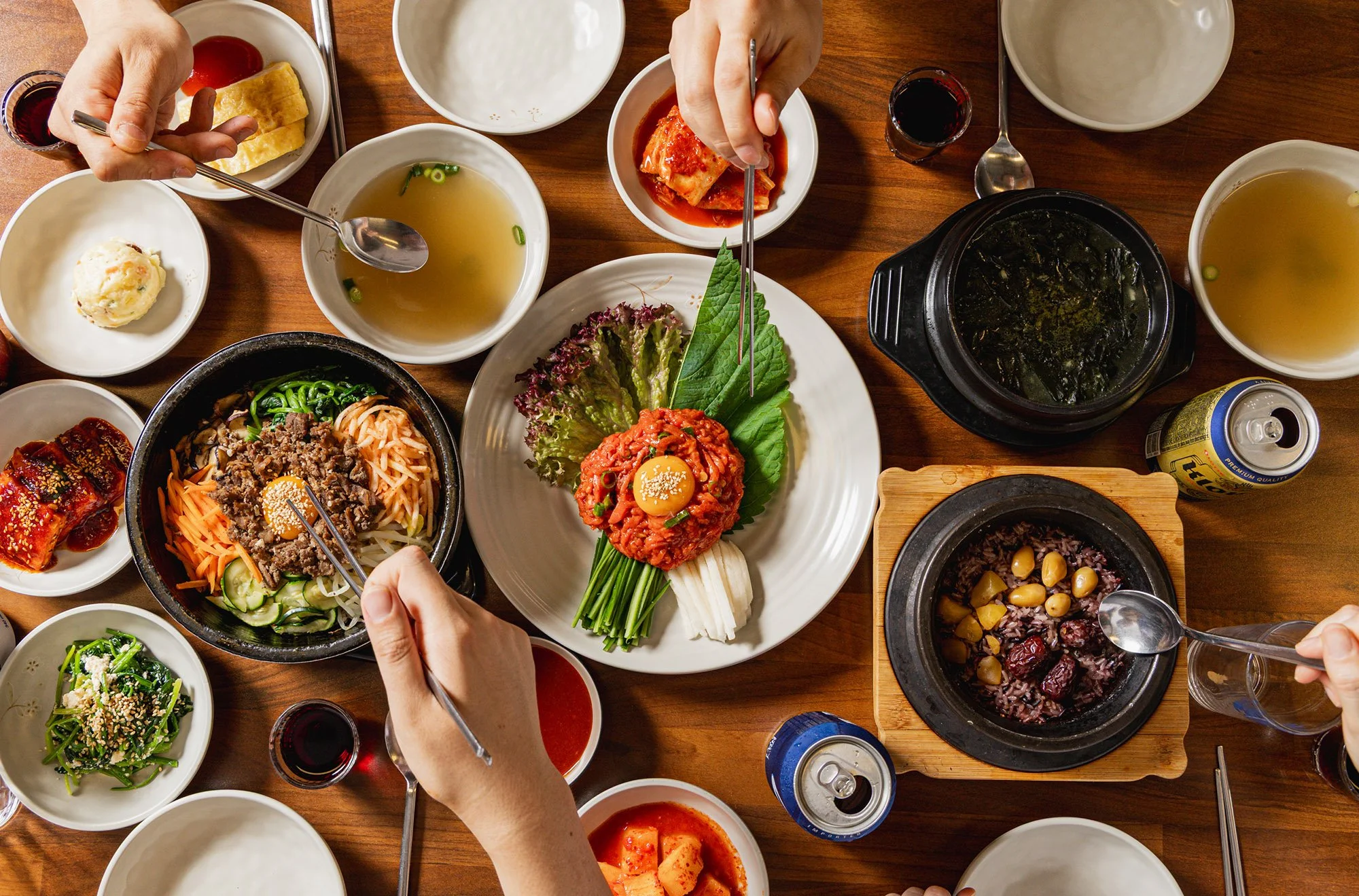 Korean dining table with various dishes including bibimbap, kimchi, soup, and side dishes, with people using chopsticks and spoons.