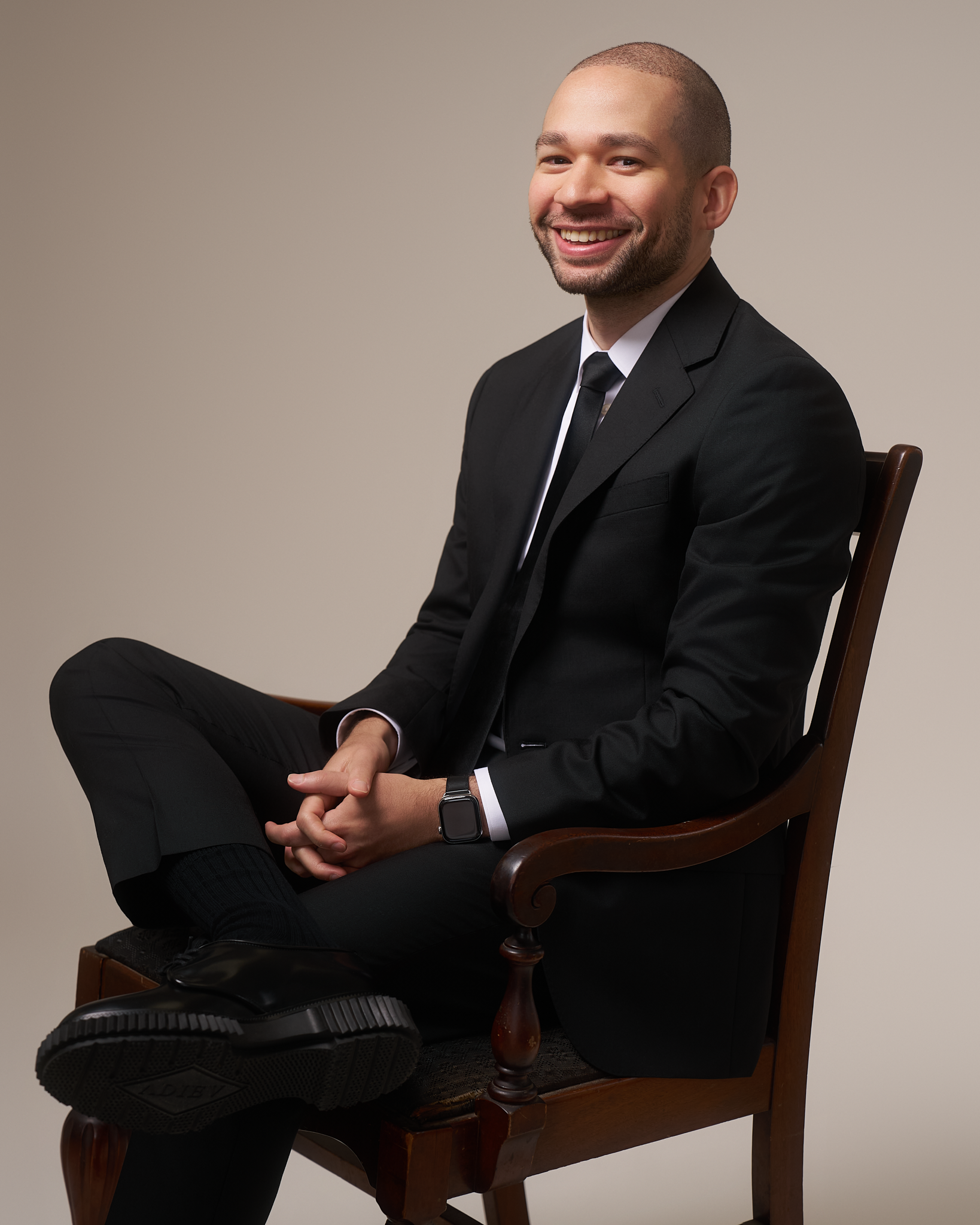 A smiling man in a black suit sitting on a wooden chair with a plain beige background.