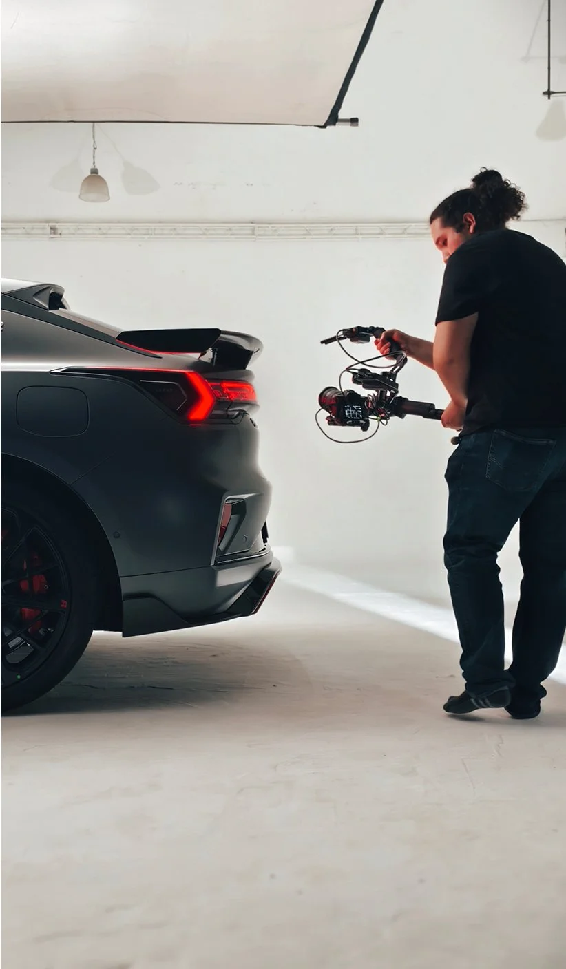 Persona en un estudio fotográfico o de grabación sosteniendo una cámara con accesorios apuntando hacia un automóvil de color gris oscuro, en un ambiente de interior.