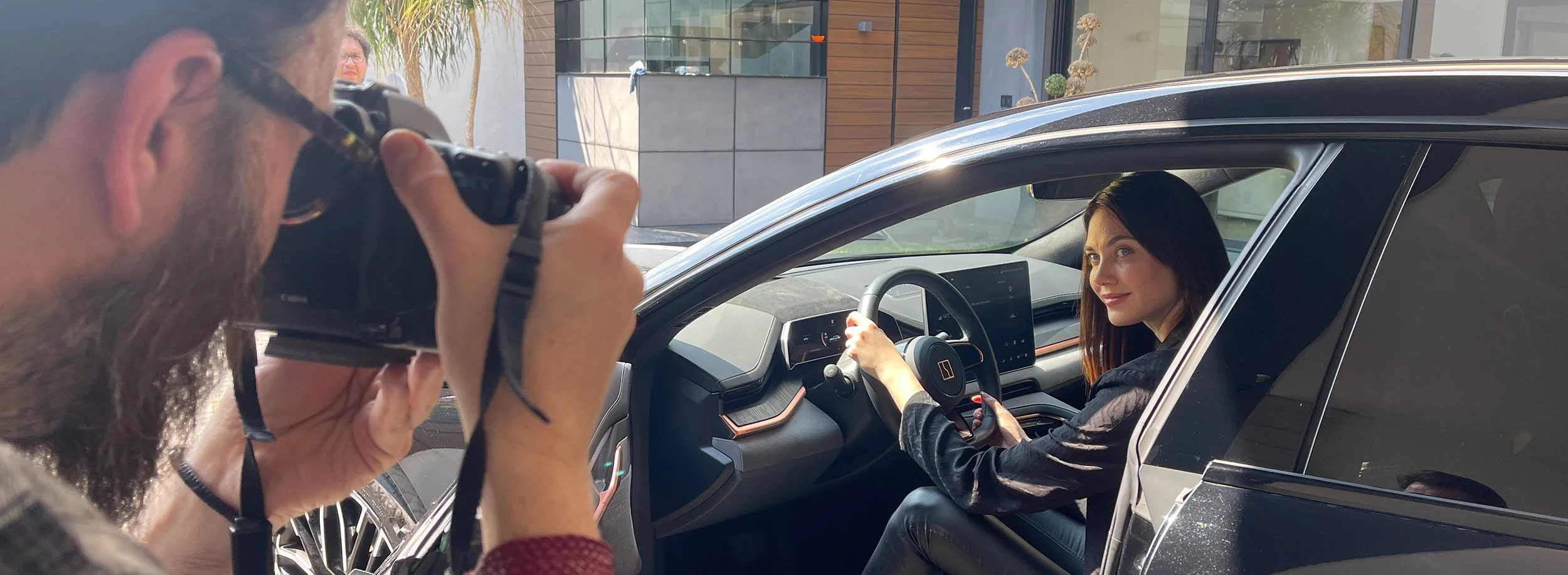 Una mujer sonriente en el asiento del conductor de un coche oscuro, posando para la cámara, mientras un hombre toma su fotografía desde afuera del vehículo