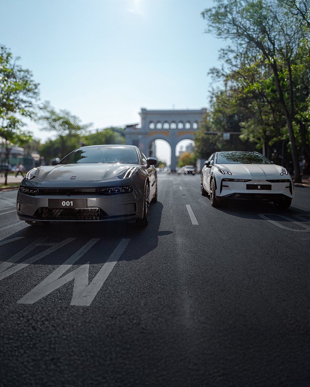 Dos autos deportivos plates de color gris y blanco estacionados en una calle con árboles y un monumento en fondo.