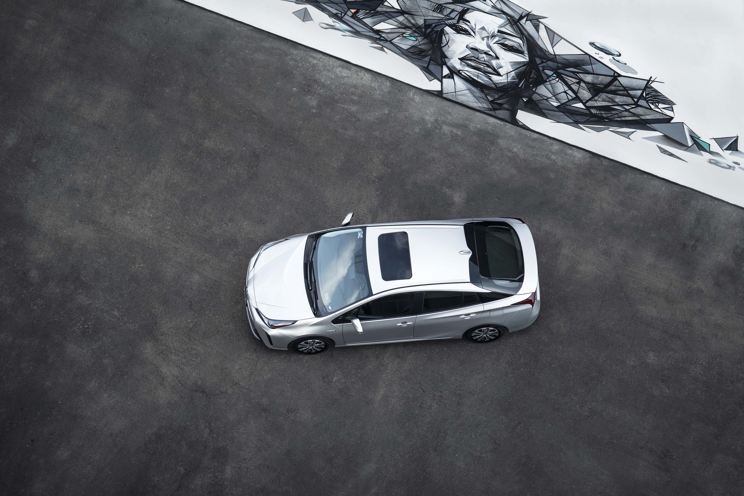 Auto plateado estacionado en una superficie plana cerca de un muro con obra de arte de rostro humano