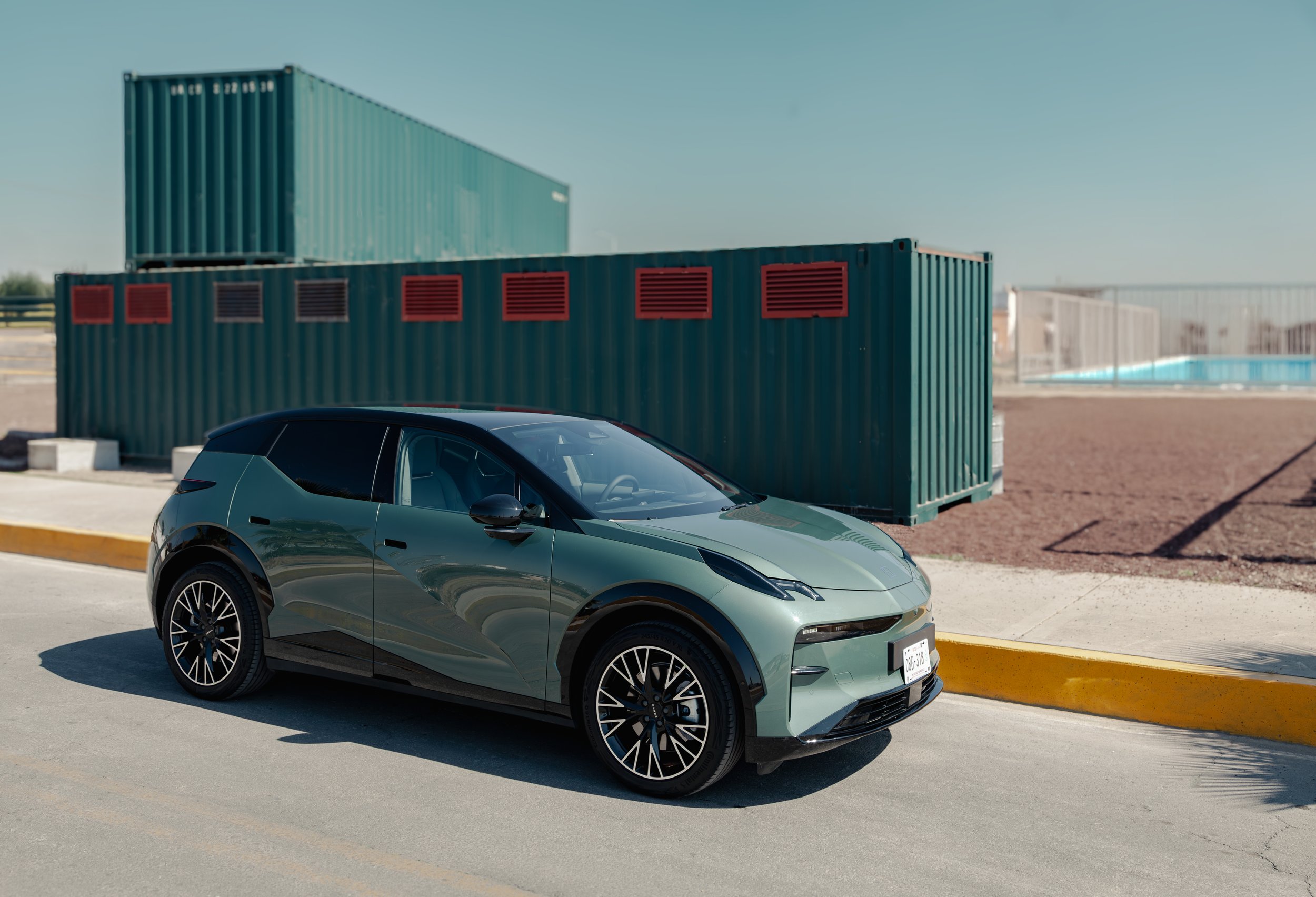 Un coche eléctrico de color gris pastel estacionado al lado de una acera en un área al aire libre, con contenedores de envío de color verde y estructuras en el fondo, bajo un cielo despejado.
