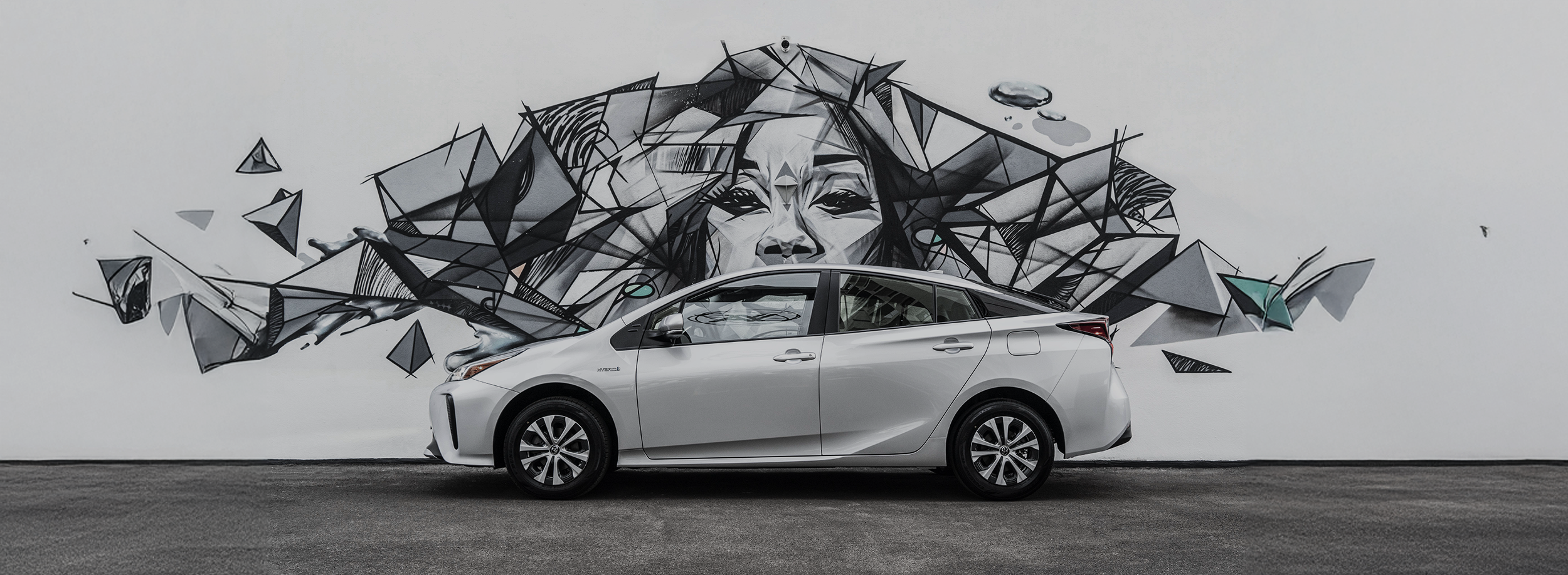 Automóvil gris estacionado frente a mural de arte en tonos grises con rostro femenino y formas geométricas.
