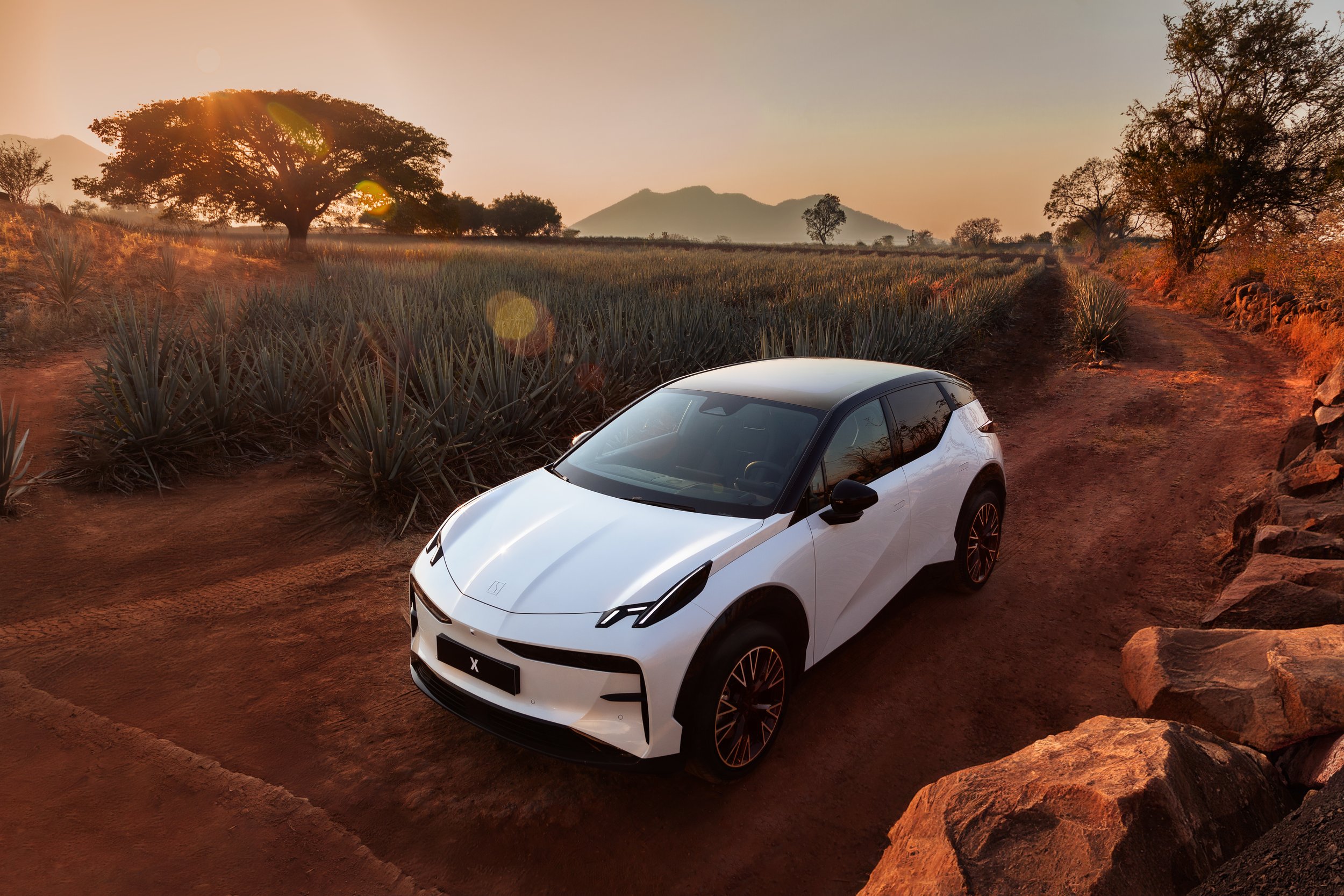 Auto eléctrico blanco en un camino de tierra en un paisaje rural con árboles y montañas al atardecer.
