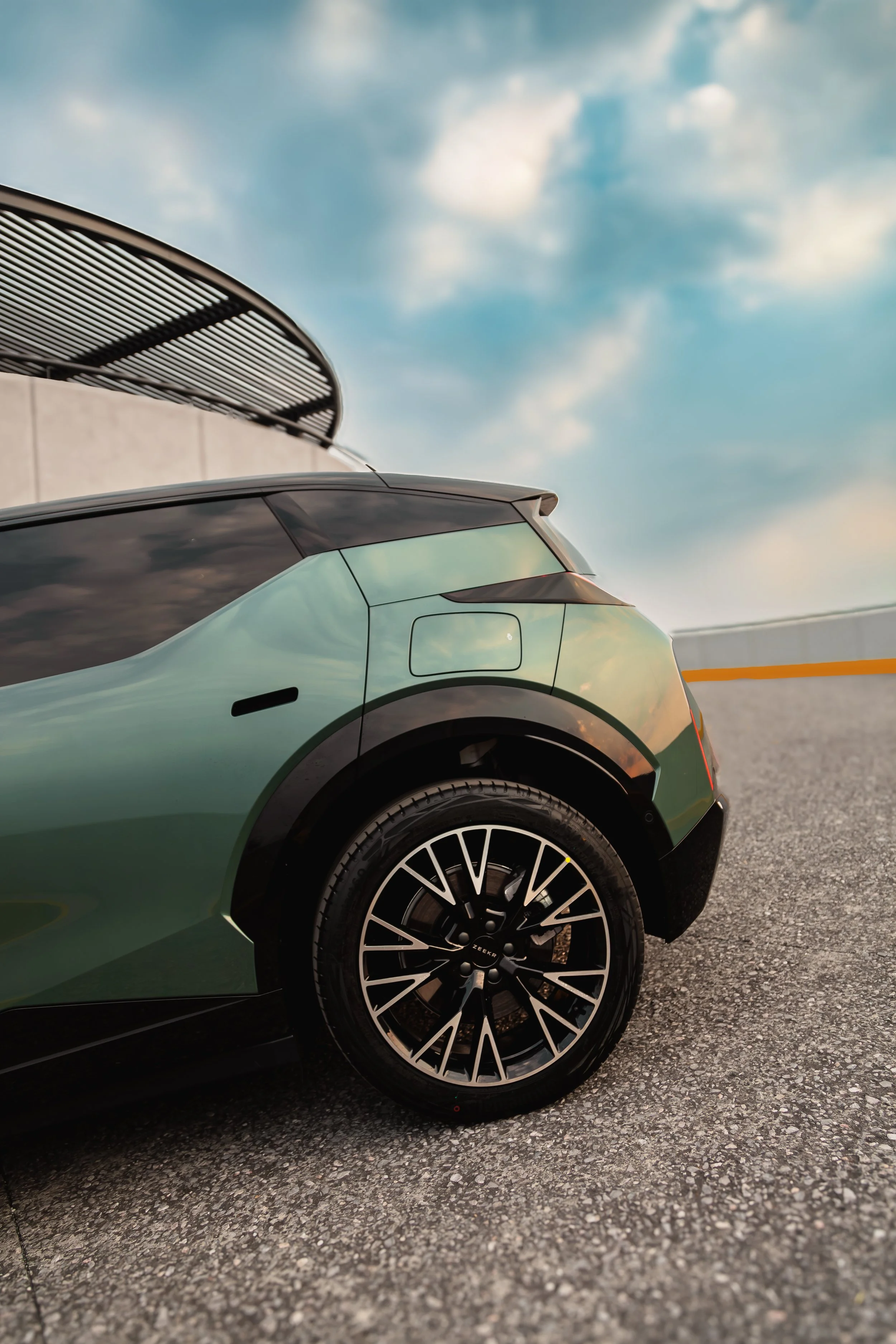 Parte trasera de un coche verde con llantas negras en un estacionamiento al aire libre, con cielo con nubes en el fondo.