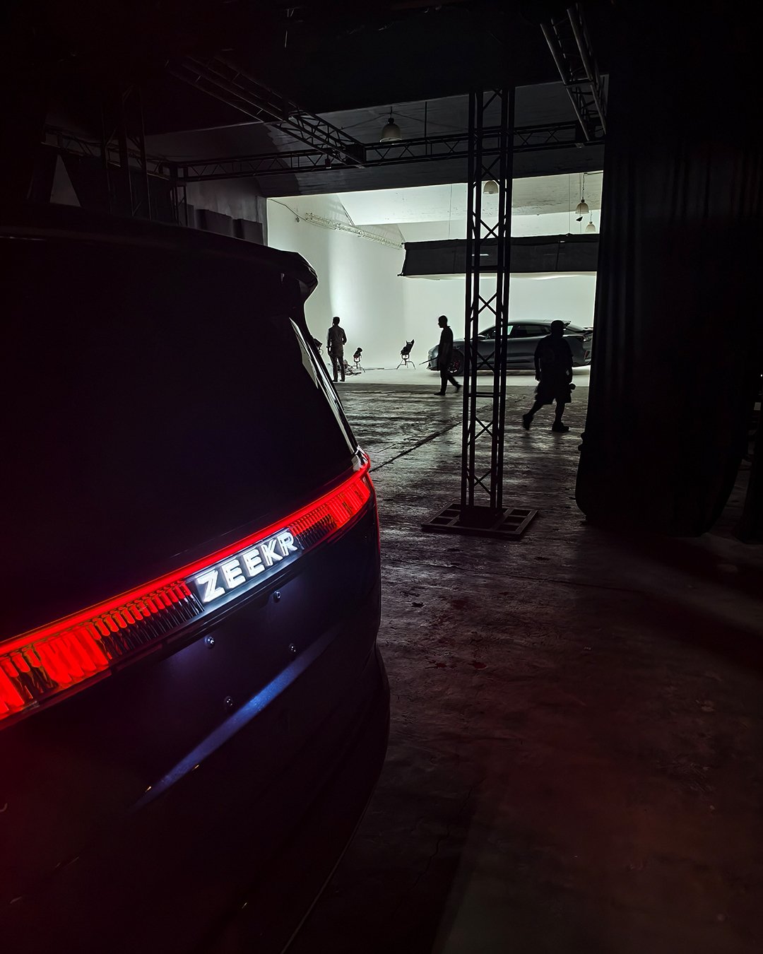 Vista desde un interior oscuro que muestra un coche negro con una línea de luces rojas en la parte trasera, y en el fondo, un set de grabación con siluetas de personas y un automóvil grande en un espacio con luz natural.