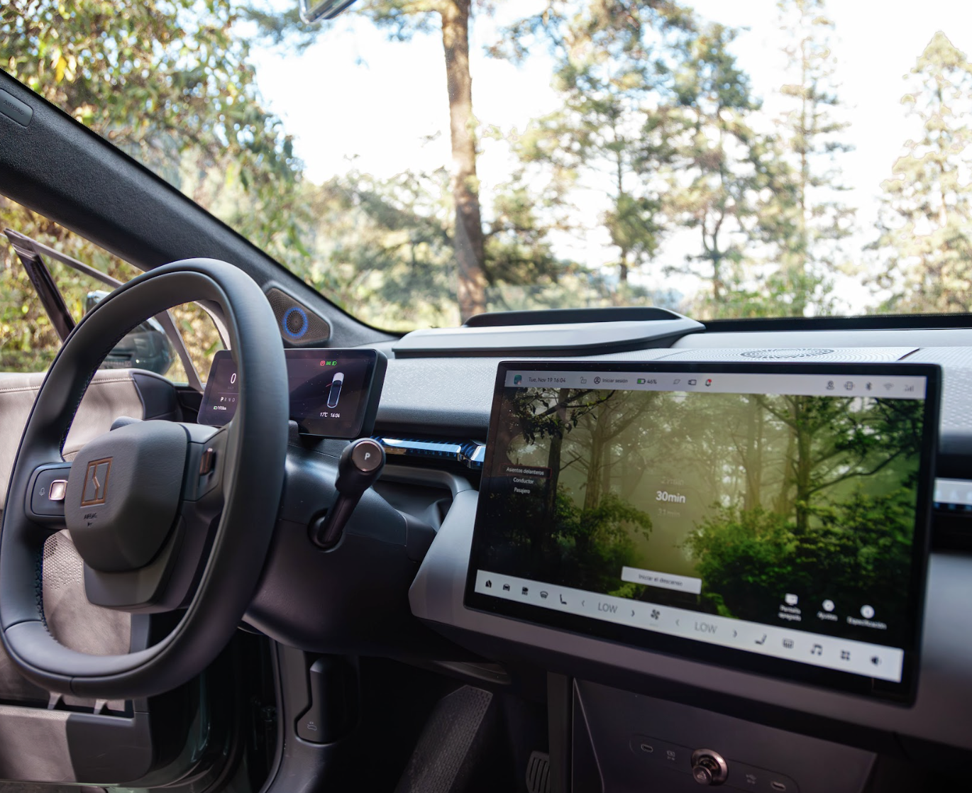 Interior de un vehículo eléctrico con panel digital y pantalla grande en medio del tablero, rodeado de naturaleza, con árboles y cielo visible a través del parabrisas.