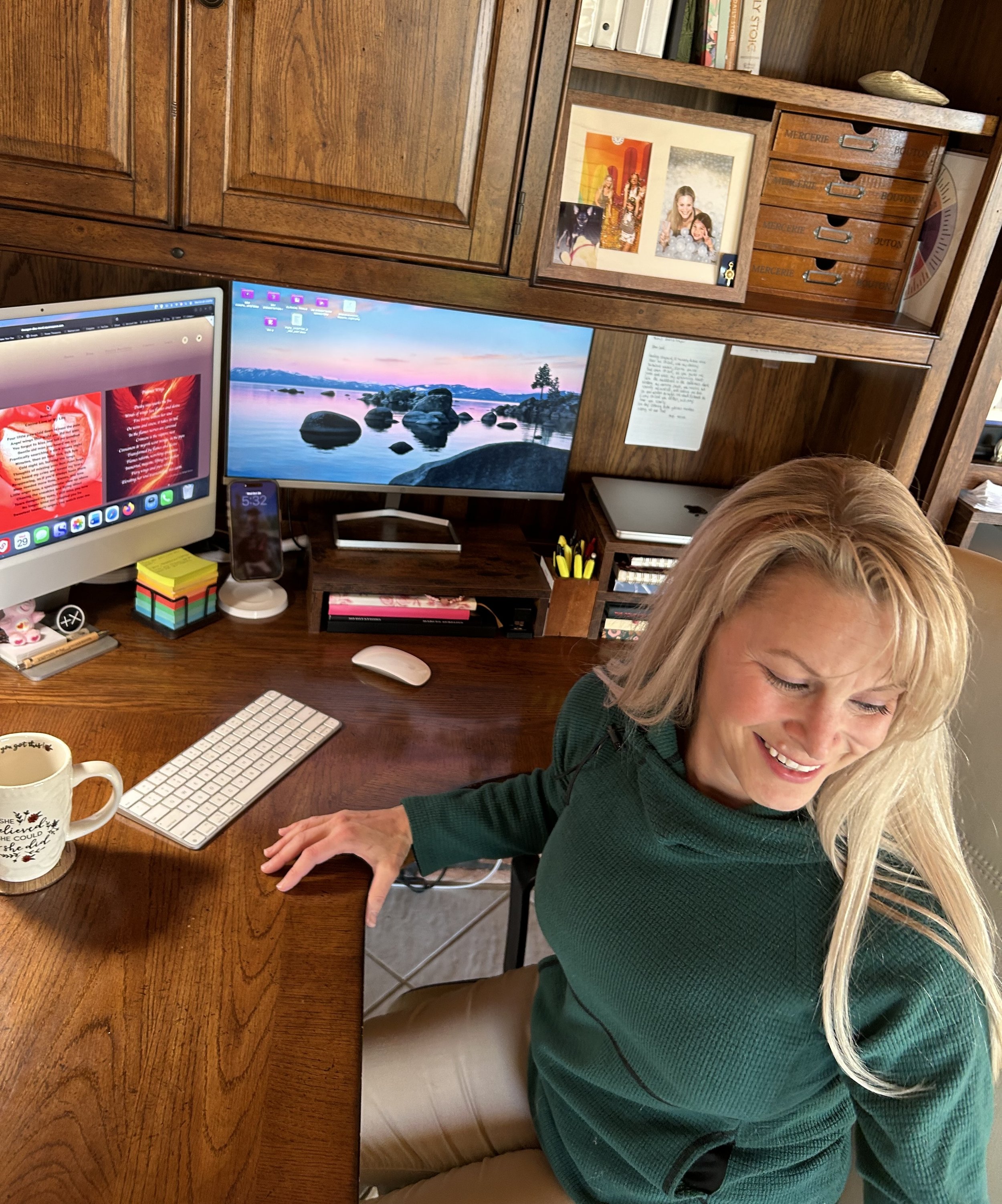 A woman with blonde hair sitting at a wooden desk, smiling and looking down, with two computer monitors, a mug, and office supplies on the desk, in a home office with wooden cabinetry and shelves behind her.