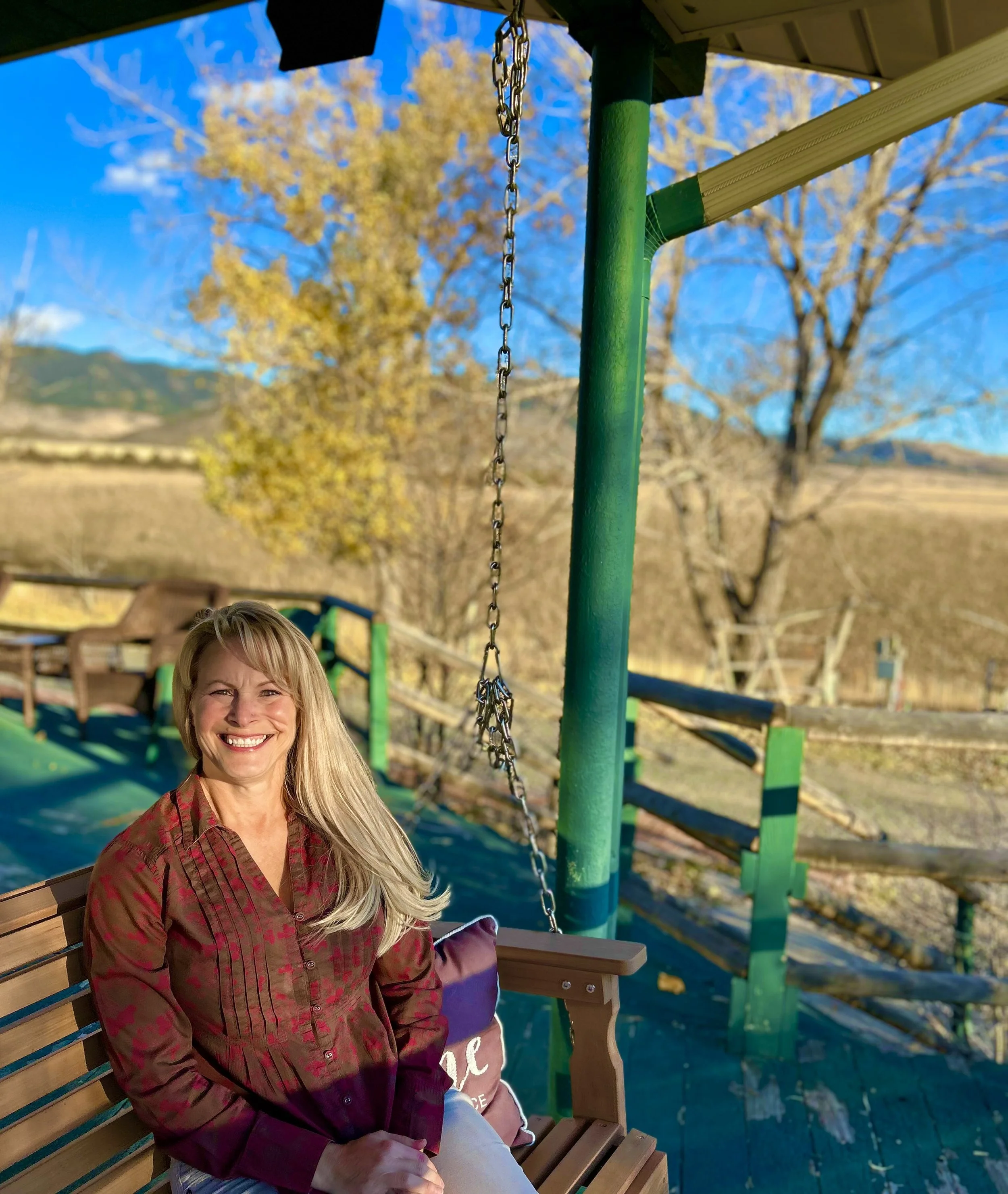 The author and creator of Gilded Inkweaver LLC has blonde hair, smiling, while sitting on a wooden swing in an outdoor park during the daytime, with a clear blue sky and trees with autumn leaves, in rural Montana.