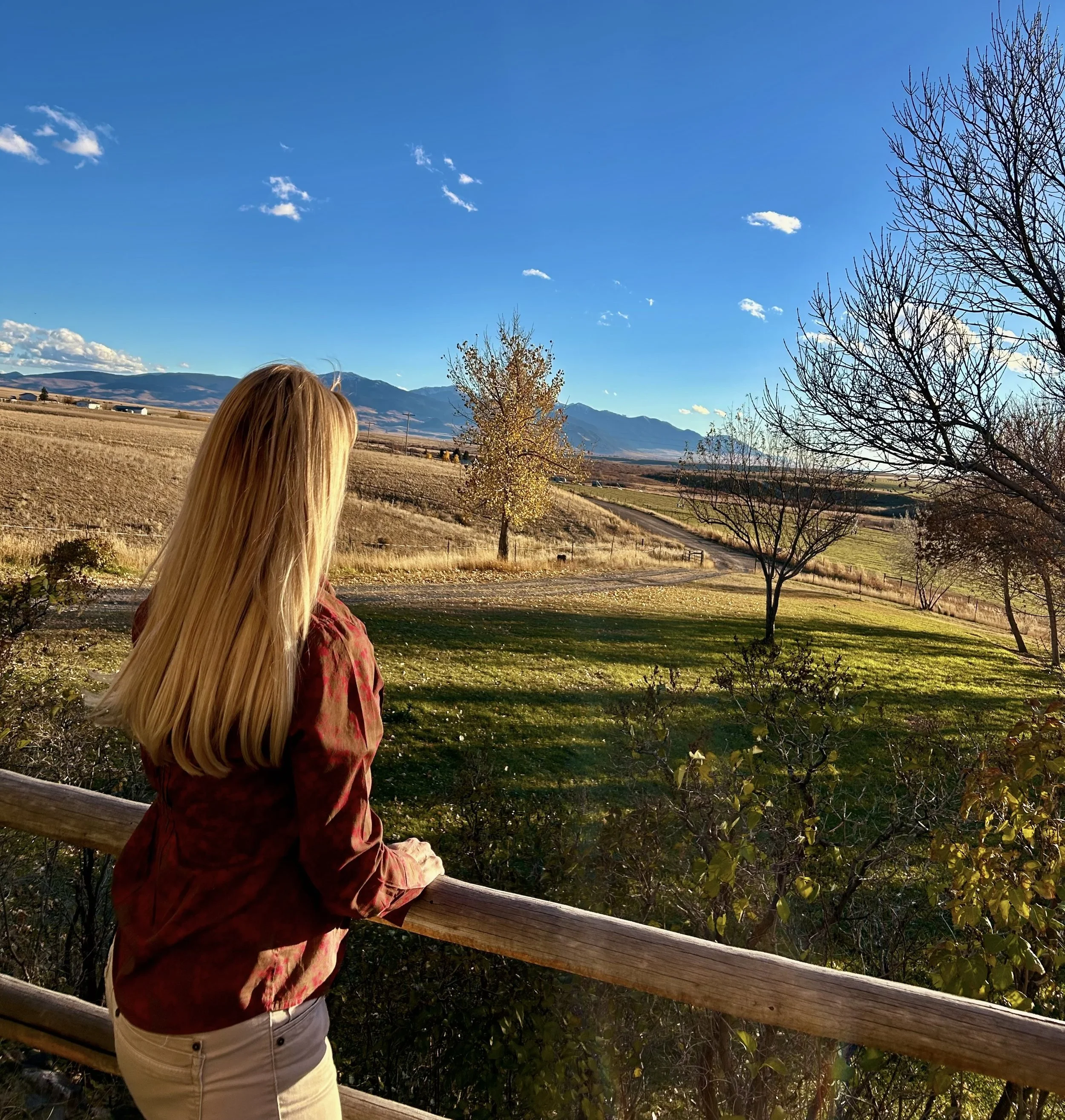 A woman with blonde hair wearing a maroon shirt and cream pants looking out over a rural landscape with trees, fields, mountains, and a clear blue sky.