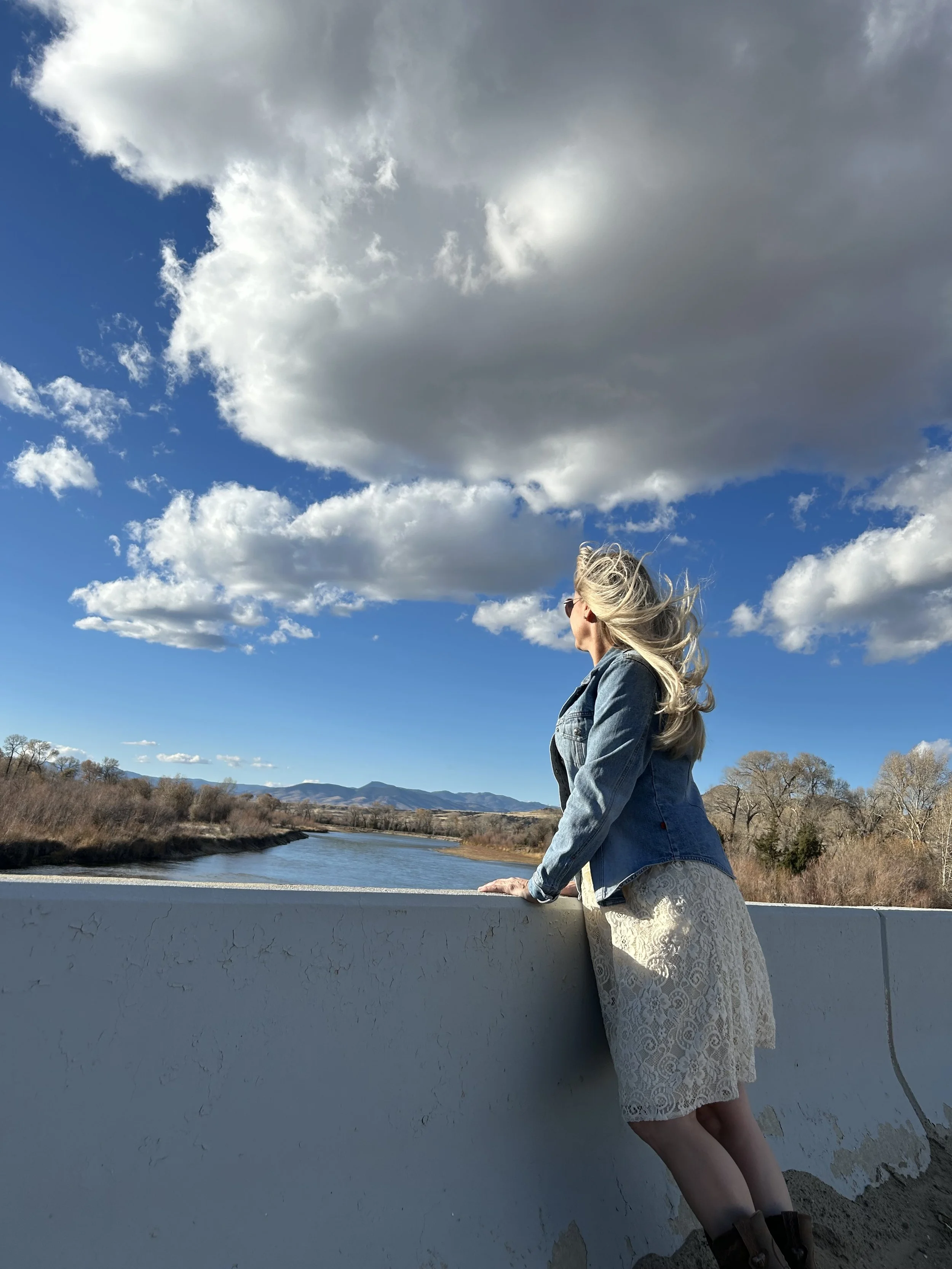 A woman with long blonde hair wearing sunglasses, a denim jacket, and a lace dress, leaning on a white concrete barrier on a bridge, looking at a river and distant mountains under a sky with large billowing clouds.