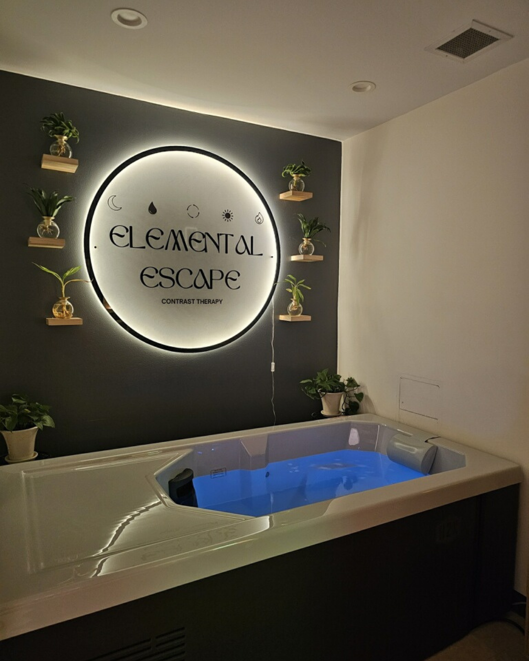 A spa room with a heated white tub filled with water, black wall with a circular sign reading 'Elemental Escape' and 'Contrast Therapy', surrounded by small potted plants on shelves and around the tub.