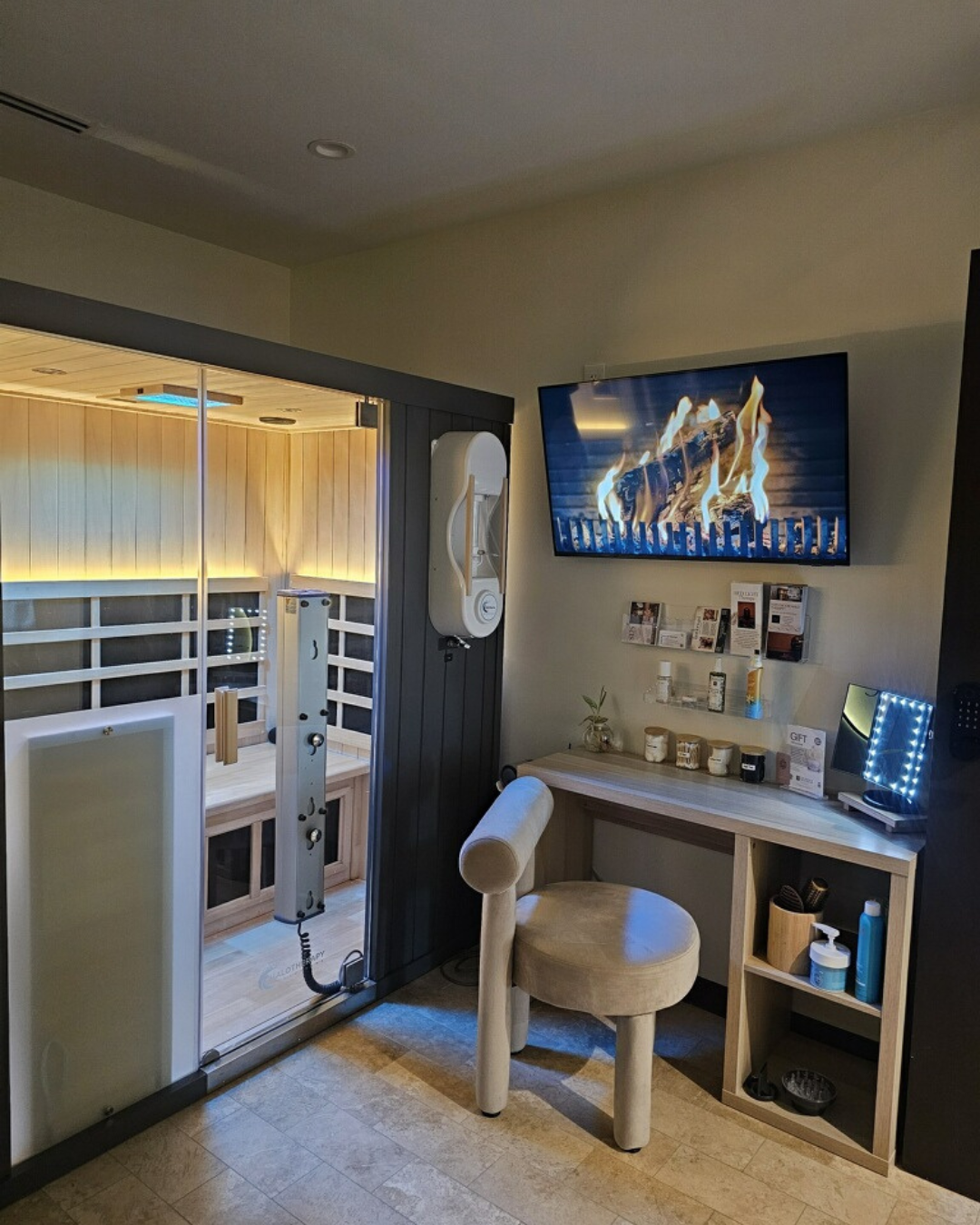 A cozy room with a mounted flat-screen TV showing a fireplace, a small wooden desk with various items, a cushioned beige chair, and a sauna with wooden interior visible through a sliding glass door.