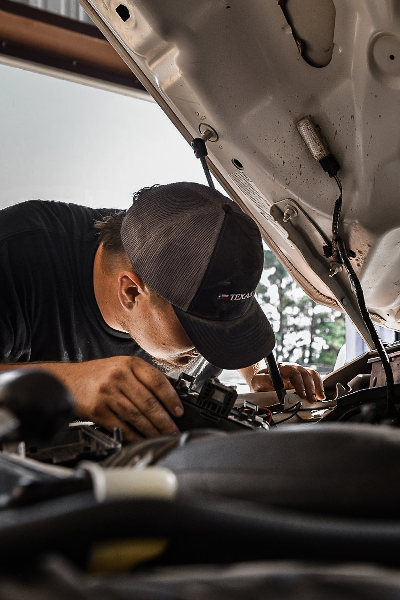 A man wearing a black baseball cap and black shirt working on a car engine with the hood open during daytime.