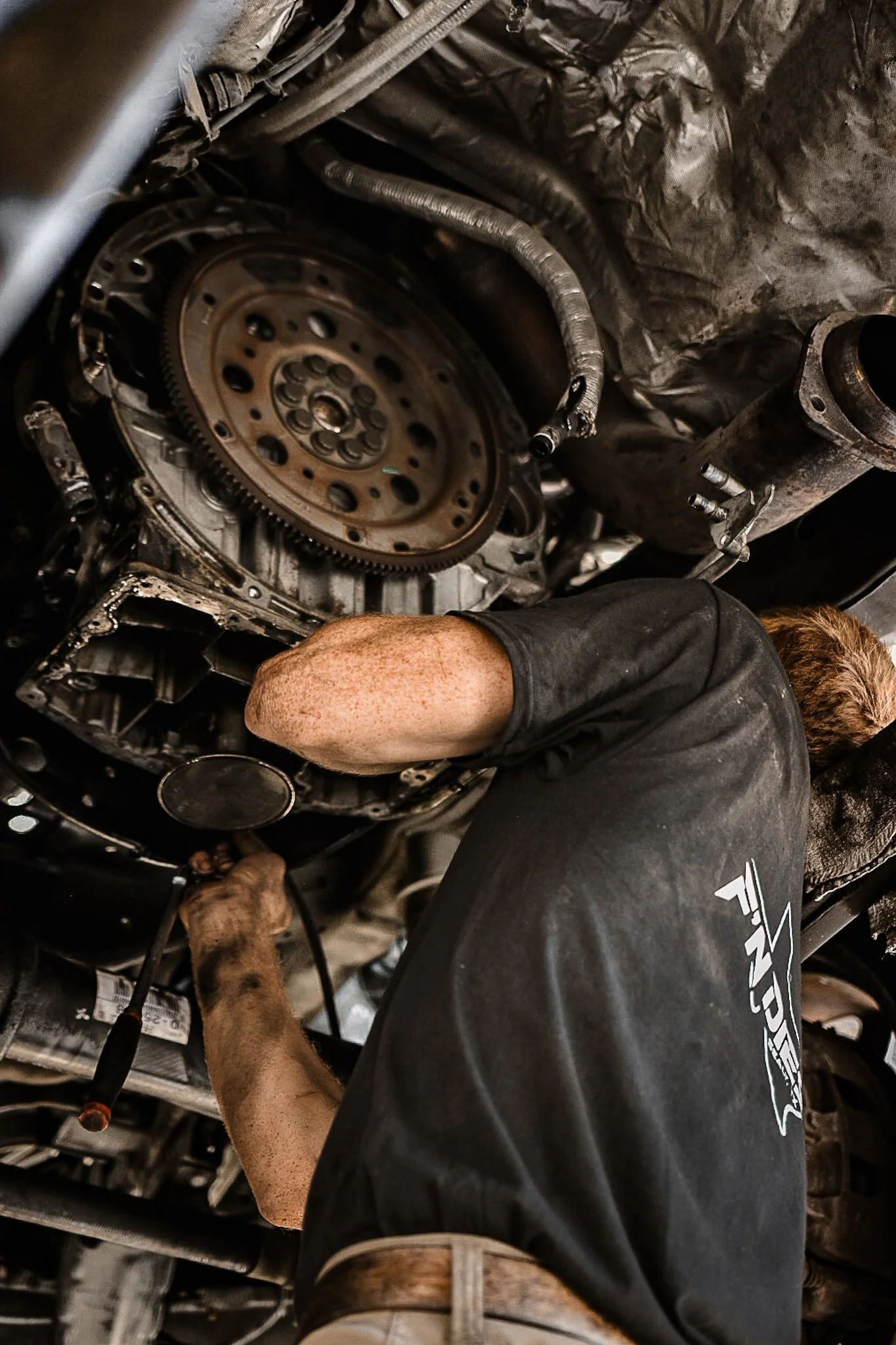A mechanic working underneath a vehicle engine, using tools to perform maintenance or repair on the engine components.