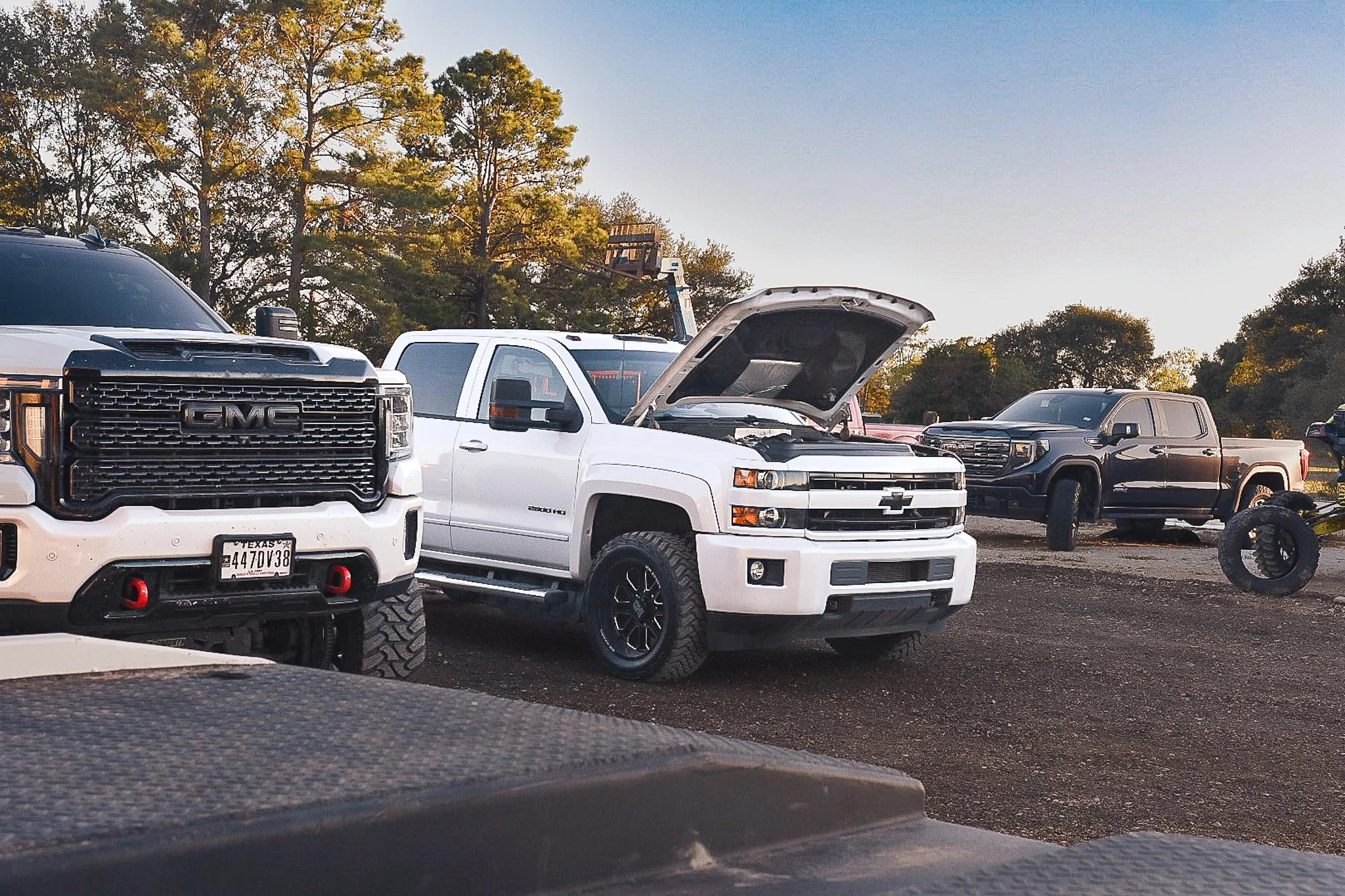 A parking lot with several pickup trucks, including a white Chevrolet with its hood open, a black GMC, and a dark gray truck in the background. There are trees and a clear sky behind the vehicles.