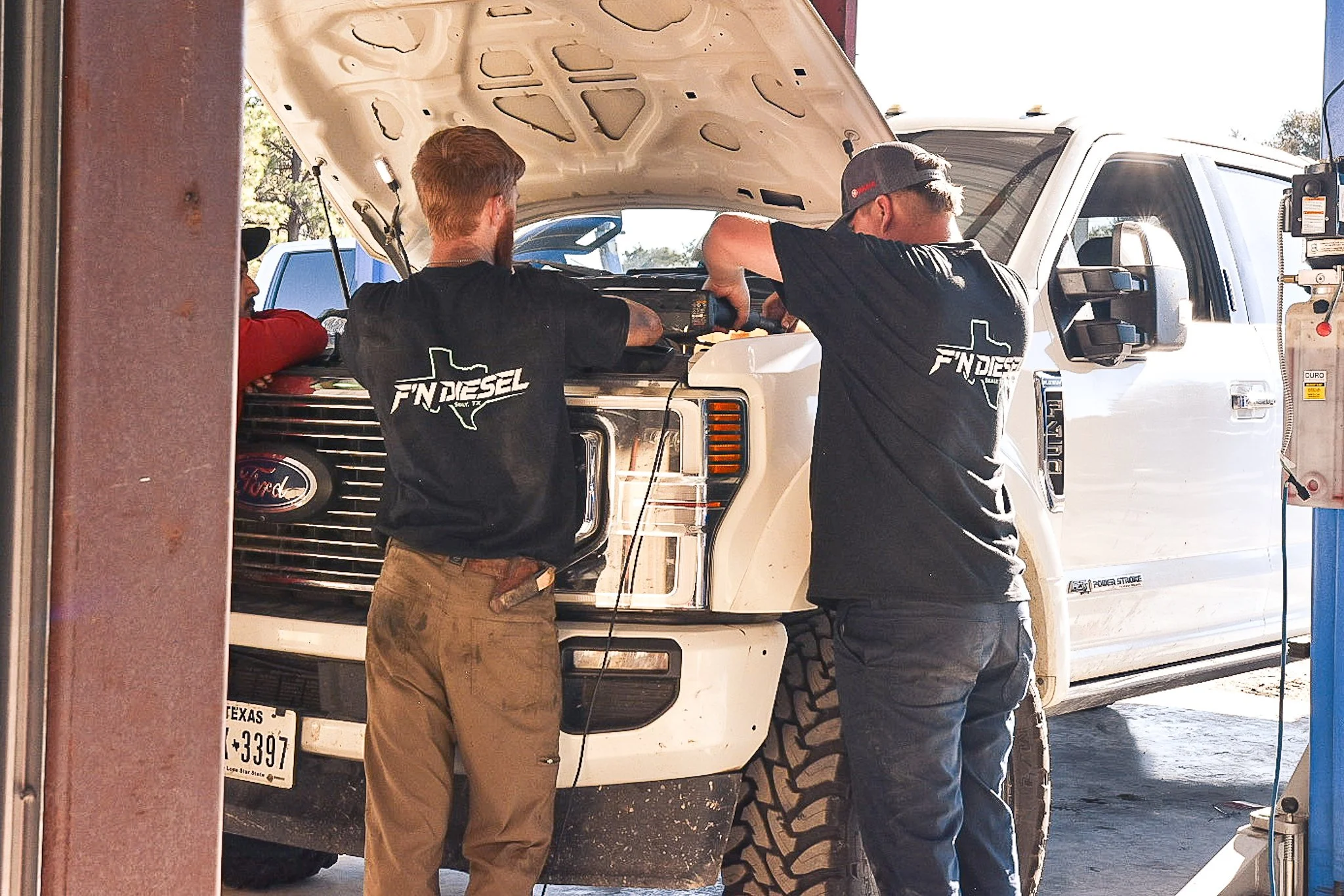 Two men working on a white Ford truck with its hood open at an auto repair shop.