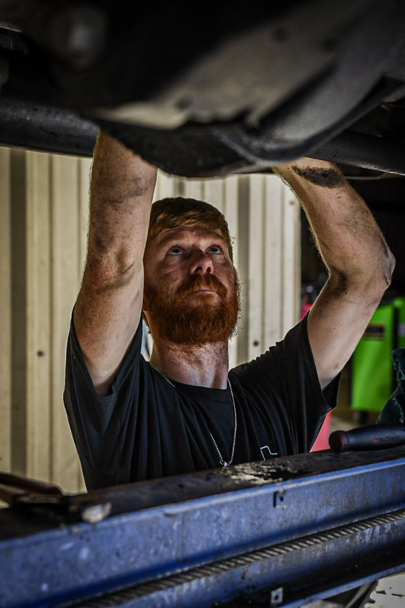 A man with a red beard and hair, wearing a black t-shirt, is working under a vehicle, inspecting or repairing it.