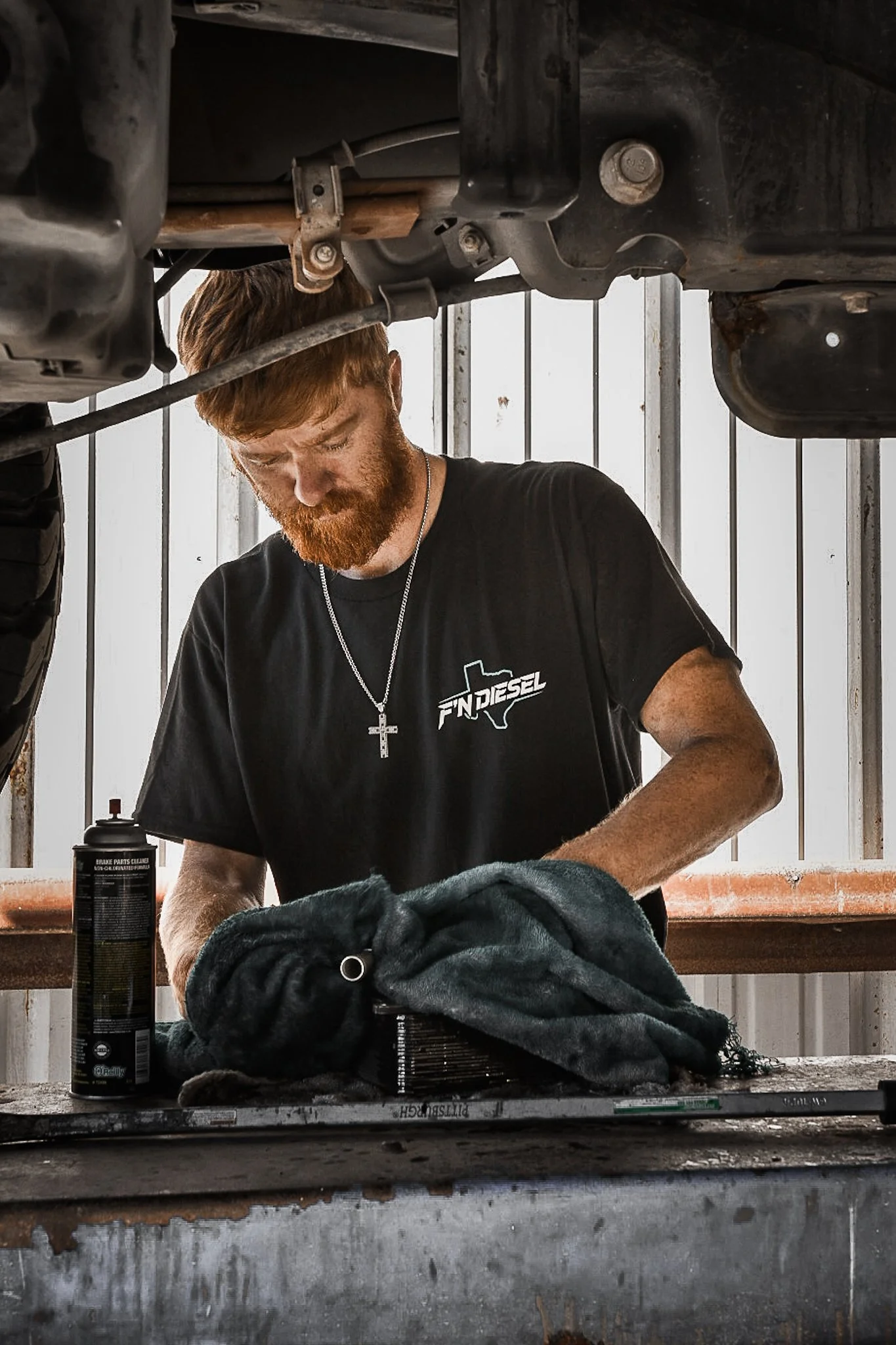 A man with reddish hair and beard wearing a black F'N Diesel t-shirt, working under a vehicle, holding a cloth or rag, with tools and spray can on the workbench