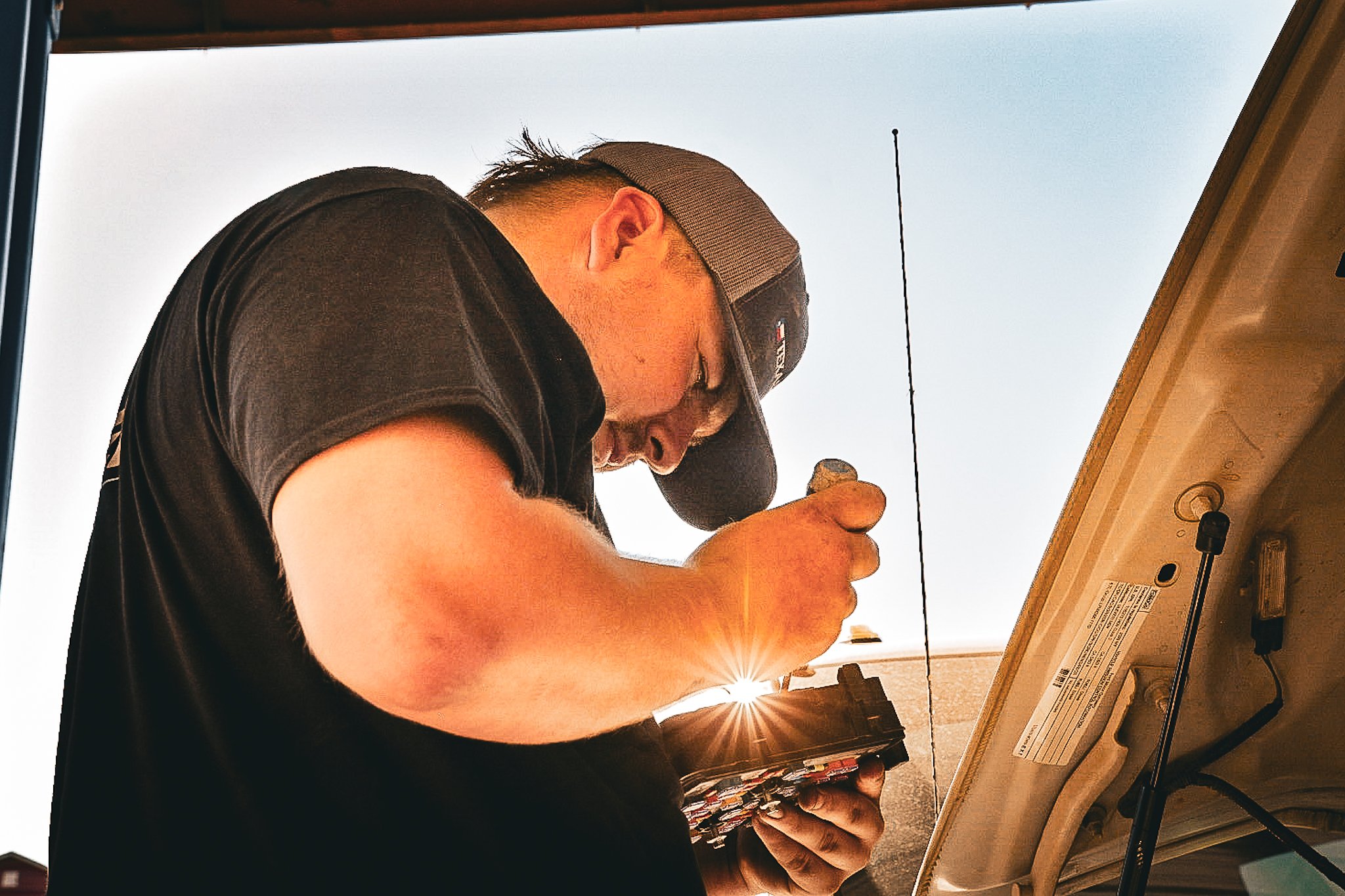 A man wearing a baseball cap and black shirt working underneath a vehicle, using a tool that emits bright sparks, with the vehicle's hood open.