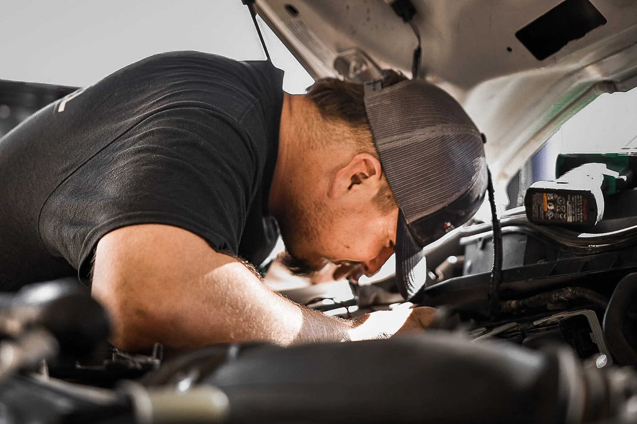 A man wearing a black t-shirt and a gray baseball cap is working under the hood of a vehicle, leaning forward with his head close to the engine.