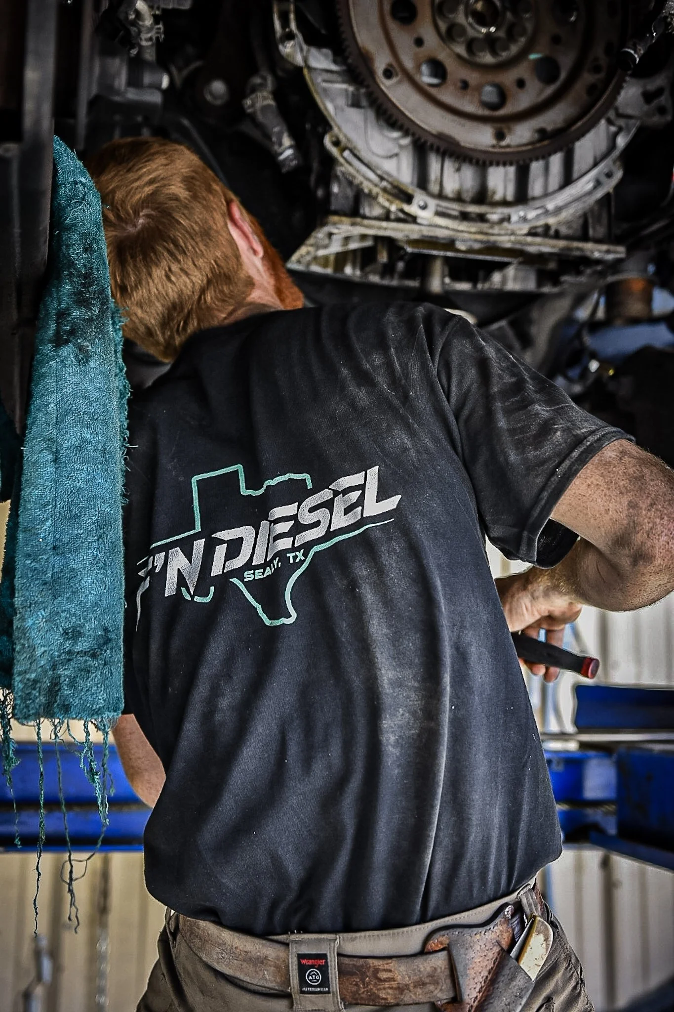 Man working underneath a vehicle, wearing a black t-shirt with a Texas outline and the words 'Indiesel Seal TX' on the back.