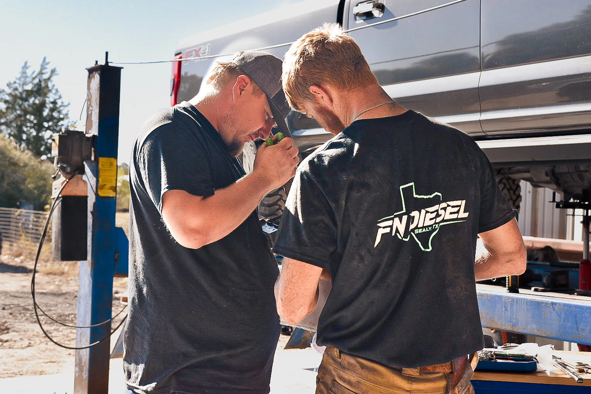 Two men working under a lifted truck in an outdoor garage, one inspecting a part, the other holding a tool, with tools on a workbench nearby.