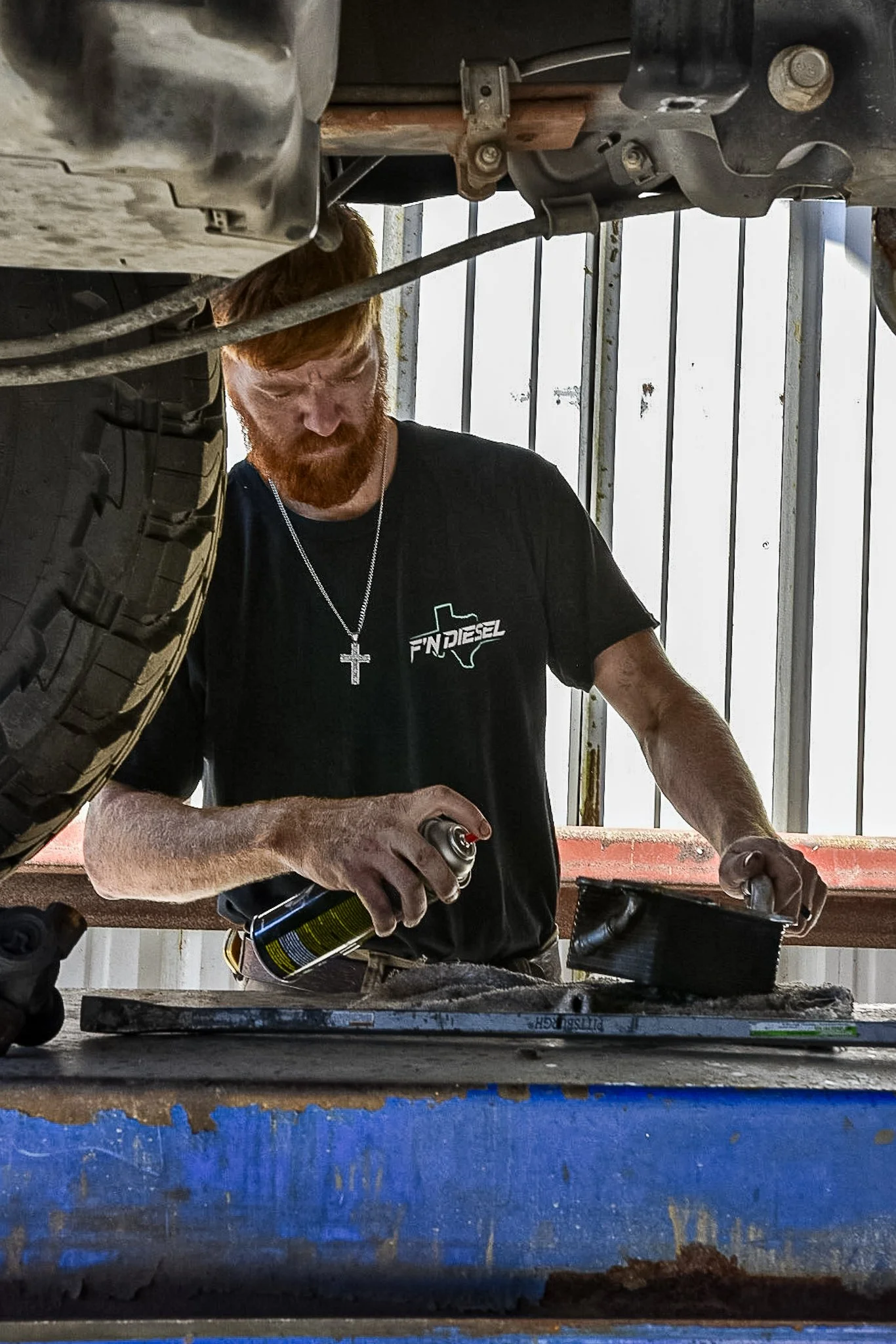 A man with a red beard wearing a black t-shirt with 'FN Diesel' logo is working underneath a vehicle, using a spray can and a filter or part in an industrial setting with metal railing in the background.