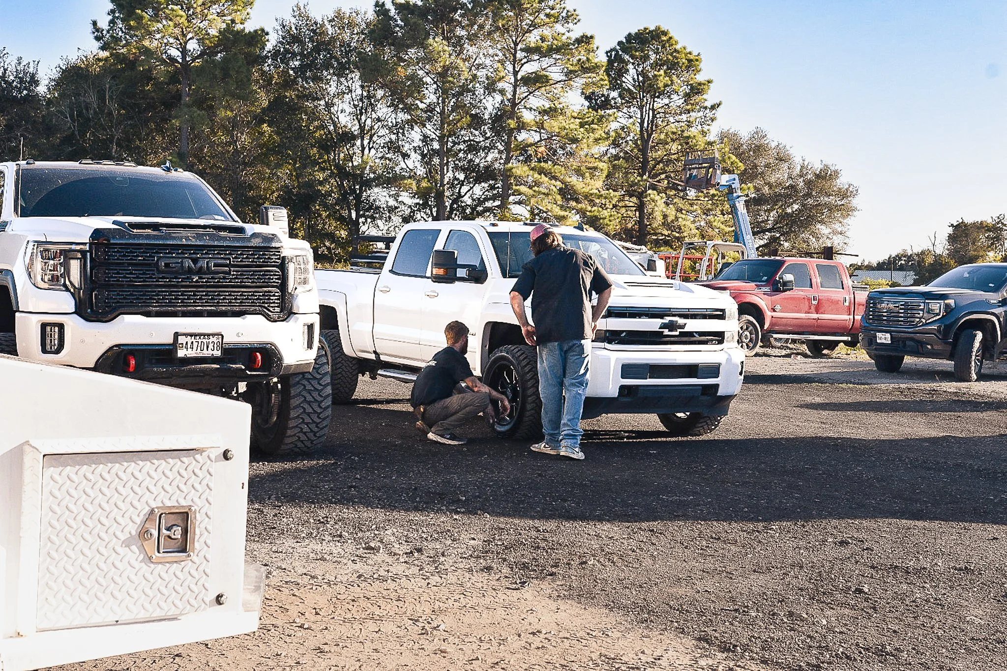 Two men inspecting the tire on a white Chevrolet Silverado pickup truck in a parking lot with several other trucks and trees in the background.