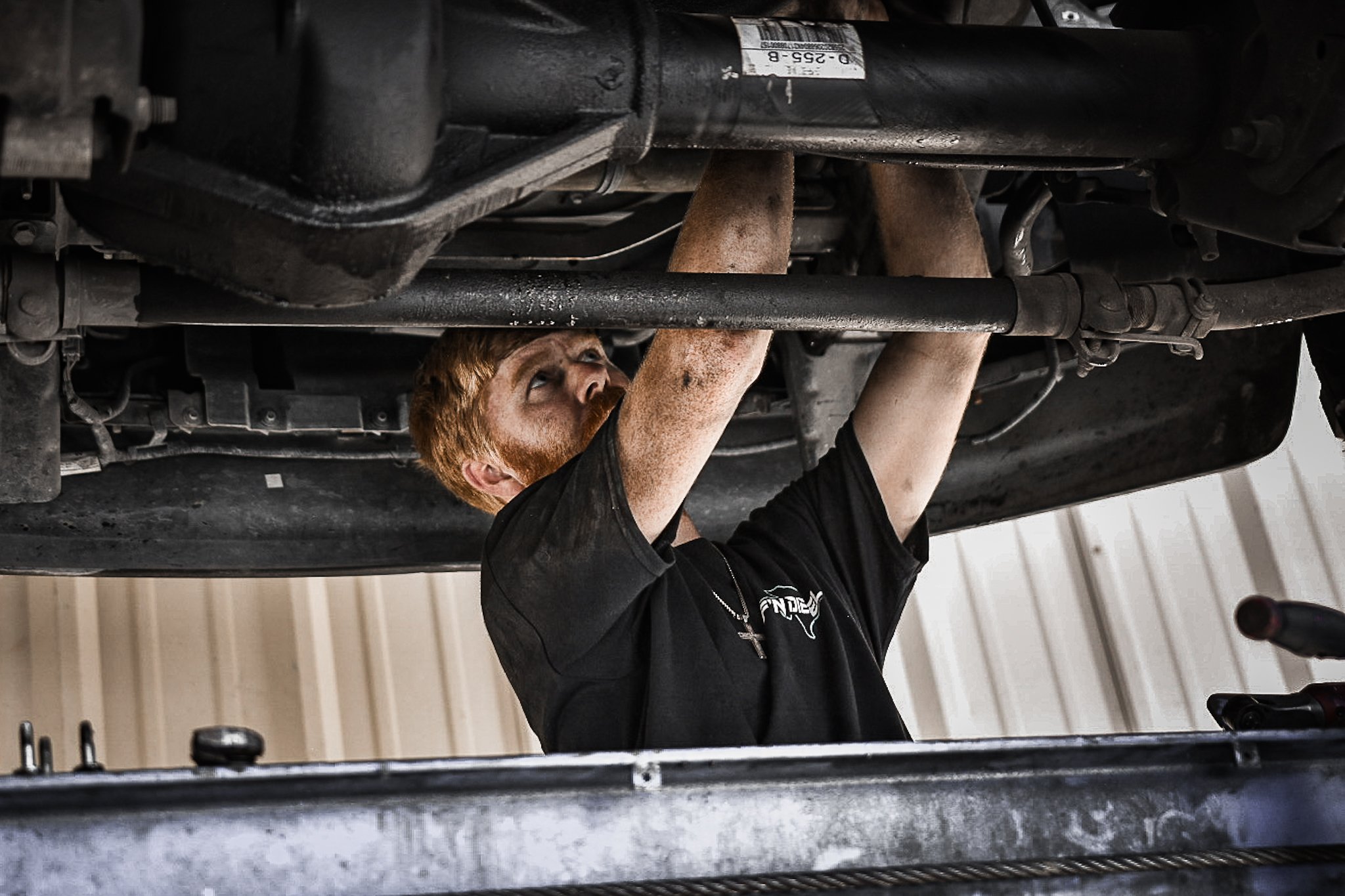 A mechanic with red hair and a black shirt working under a car, inspecting or repairing the underside.