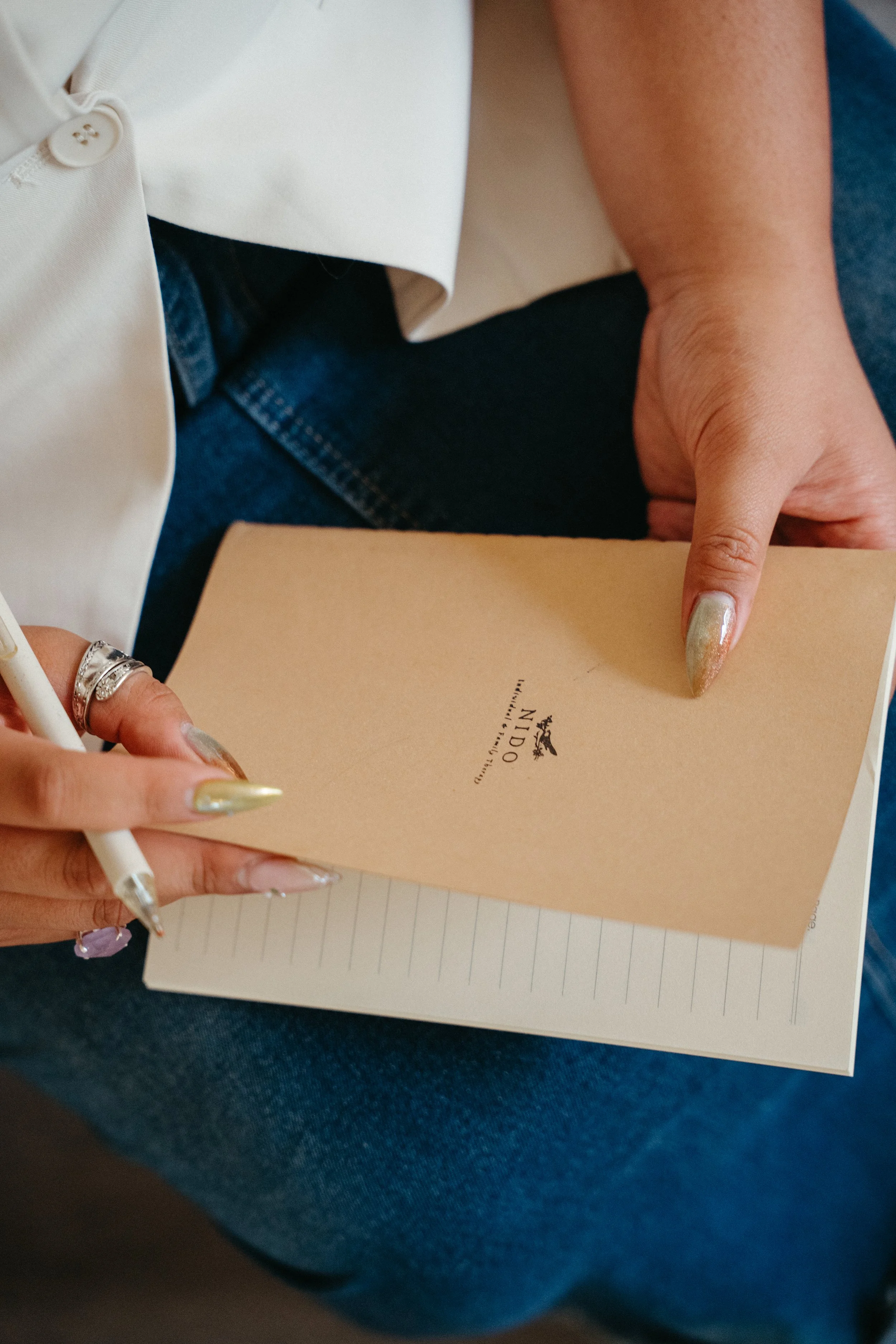 Photo of hands with mermaid colored nails and rings holding a pen and notebook that says Nido in their lap