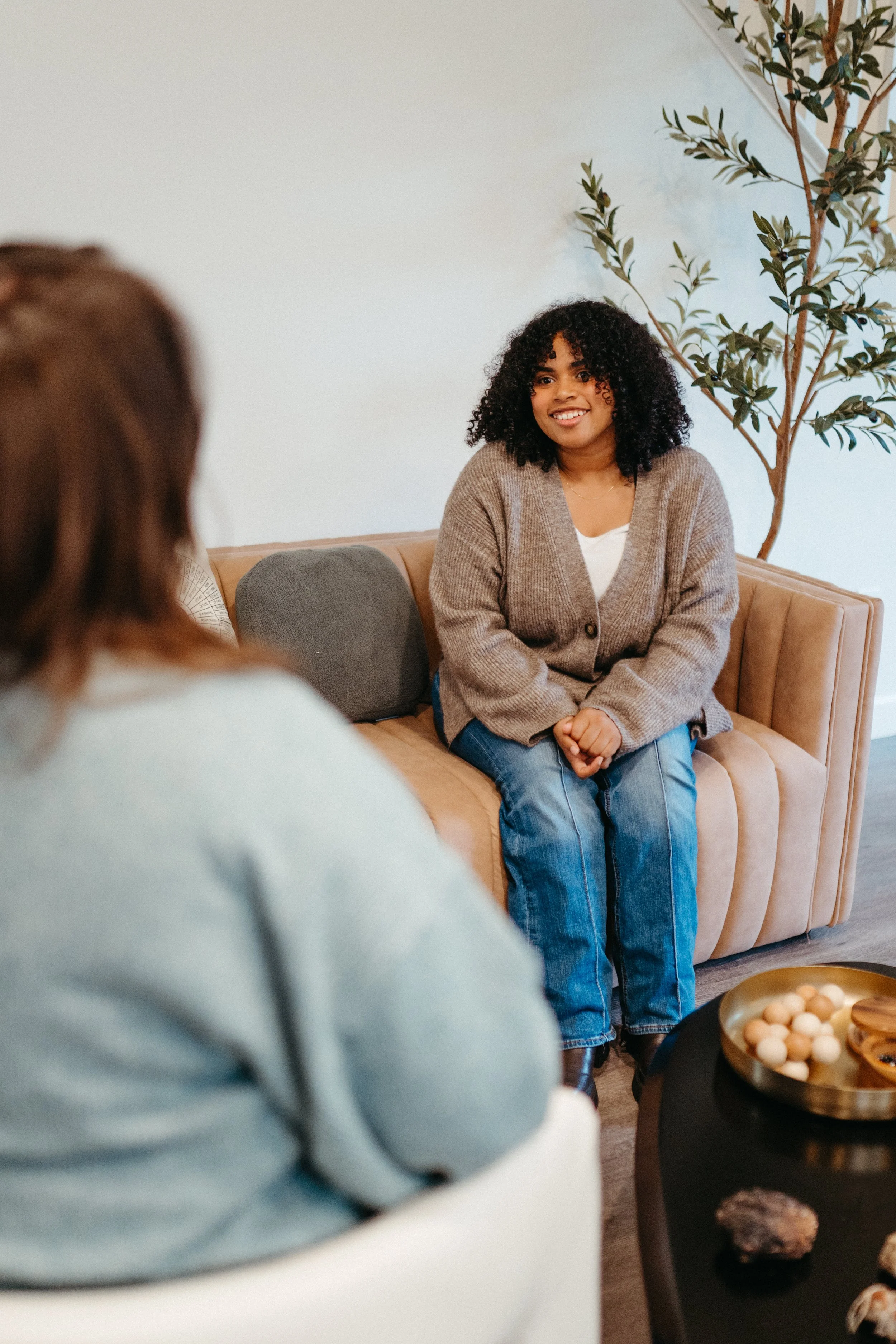 Photo of a smiling curly haired client wearing a cozy sweater sitting on a couch facing their therapist