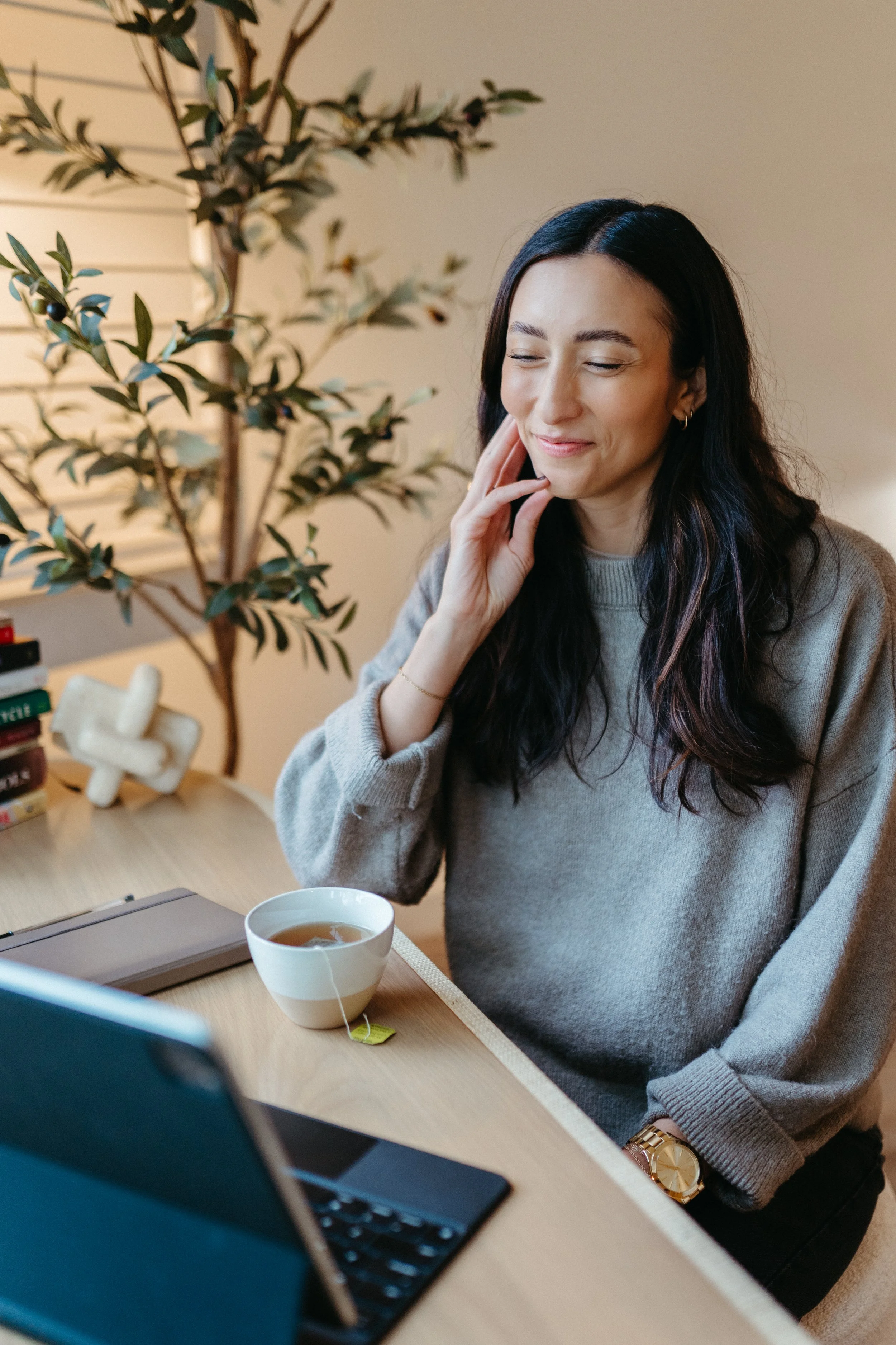 Photo of Nido IFT founder Sarah Finch sitting at a desk with a cup of tea and smiling on a telehealth session