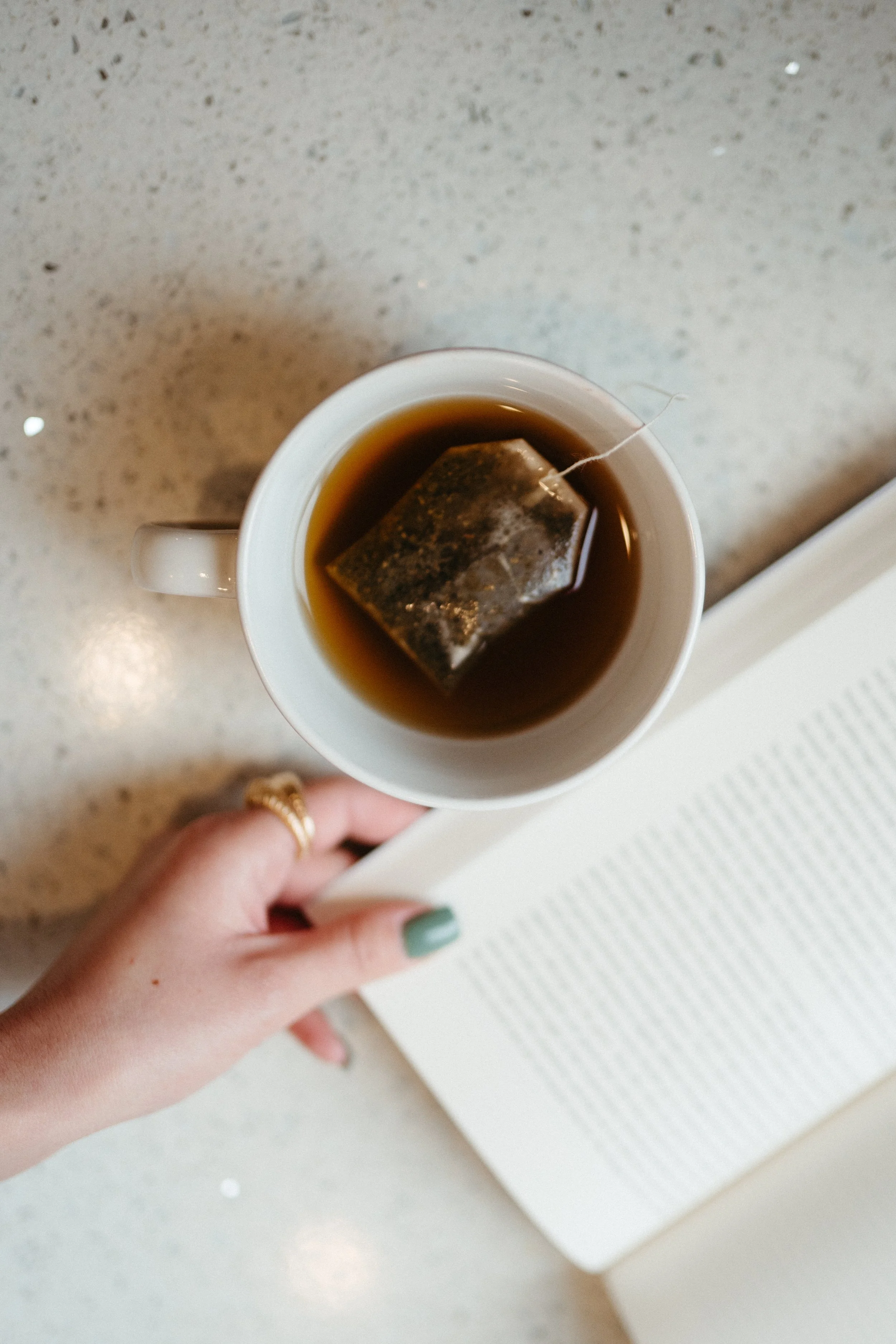 Photo from above of a teacup and a hand with green nails and a gold ring holding the edge of a book