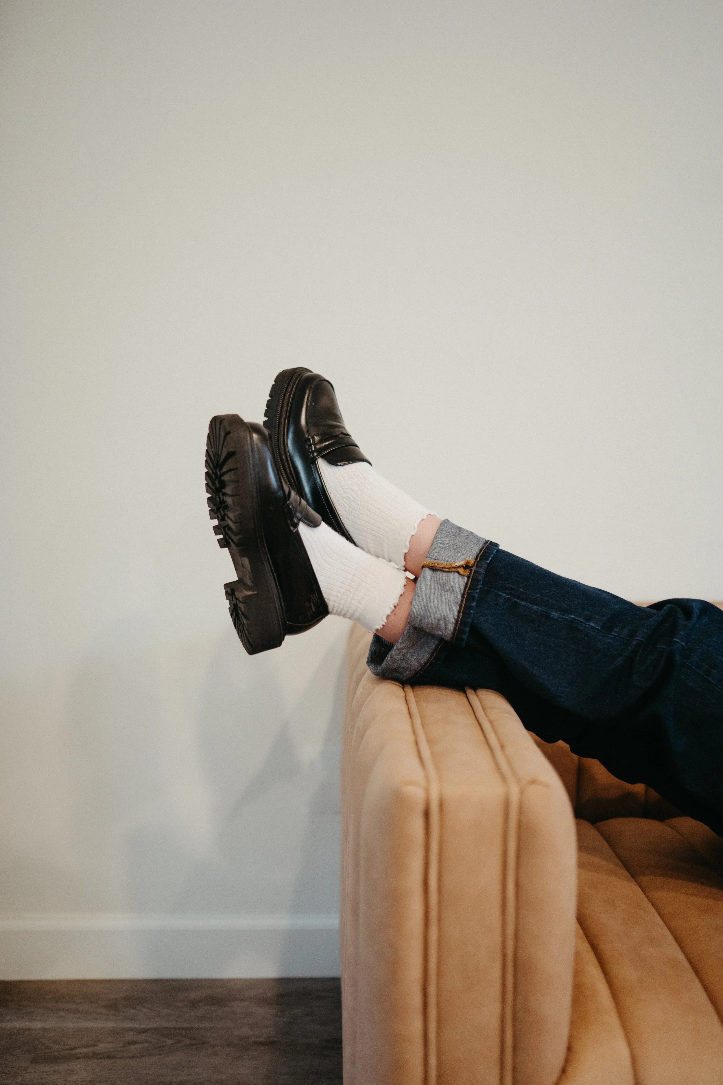 Photo of lower legs wearing black oxford shoes and white socks propped up casually on the arm of a couch